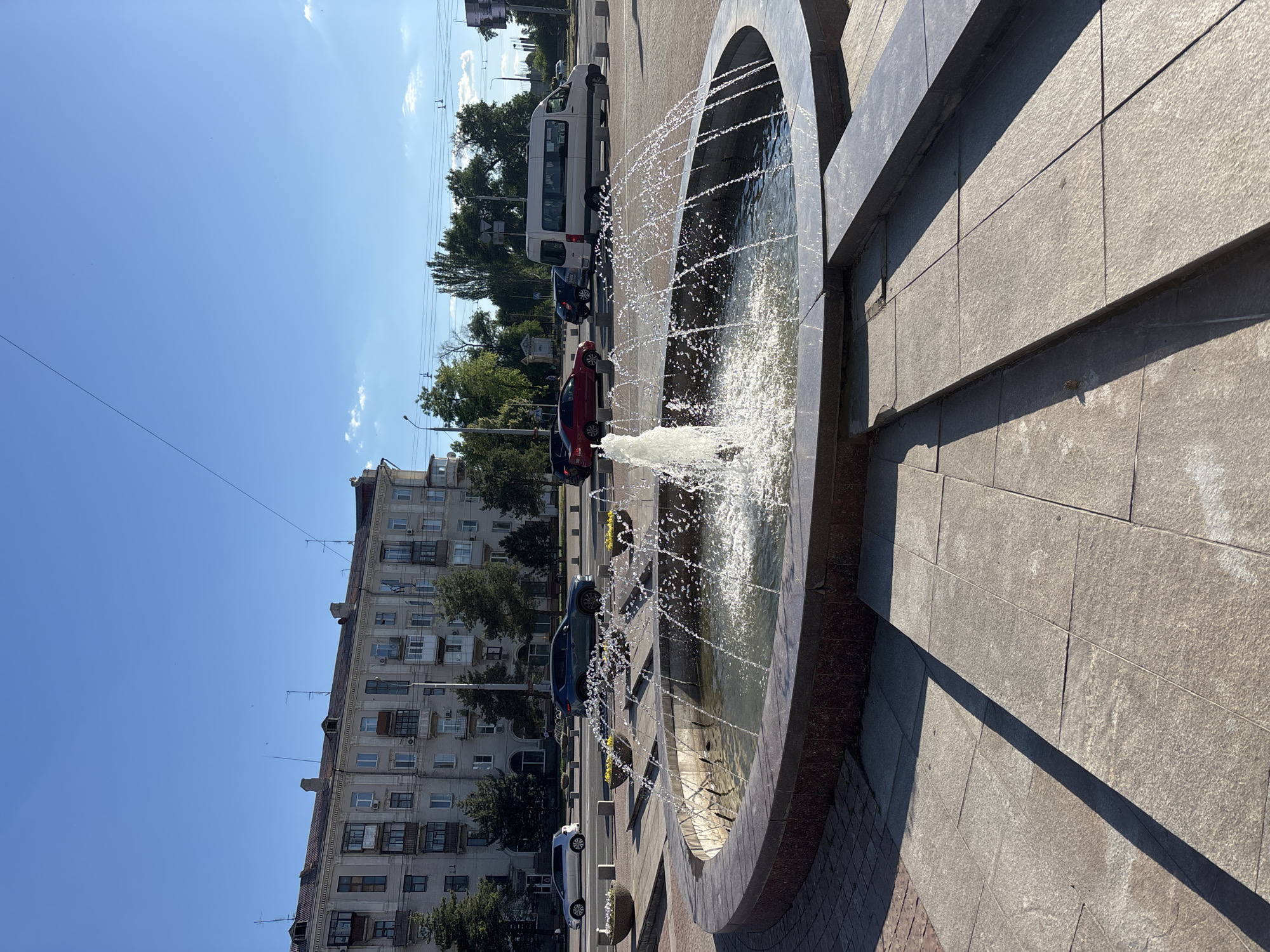 A bright, sunny day with a clear blue sky over a city street. In the foreground, a circular fountain with water spraying upwards is set in a paved plaza with stone steps. The fountain has a dark stone border. Beyond the fountain, cars are driving on a multi-lane road. On the left side of the road, there is a light-colored multi-story apartment building with many windows and balconies. Trees line the street between the road and the building. On the right side of the road, there are more trees and a white van. The overall scene depicts a typical urban environment.