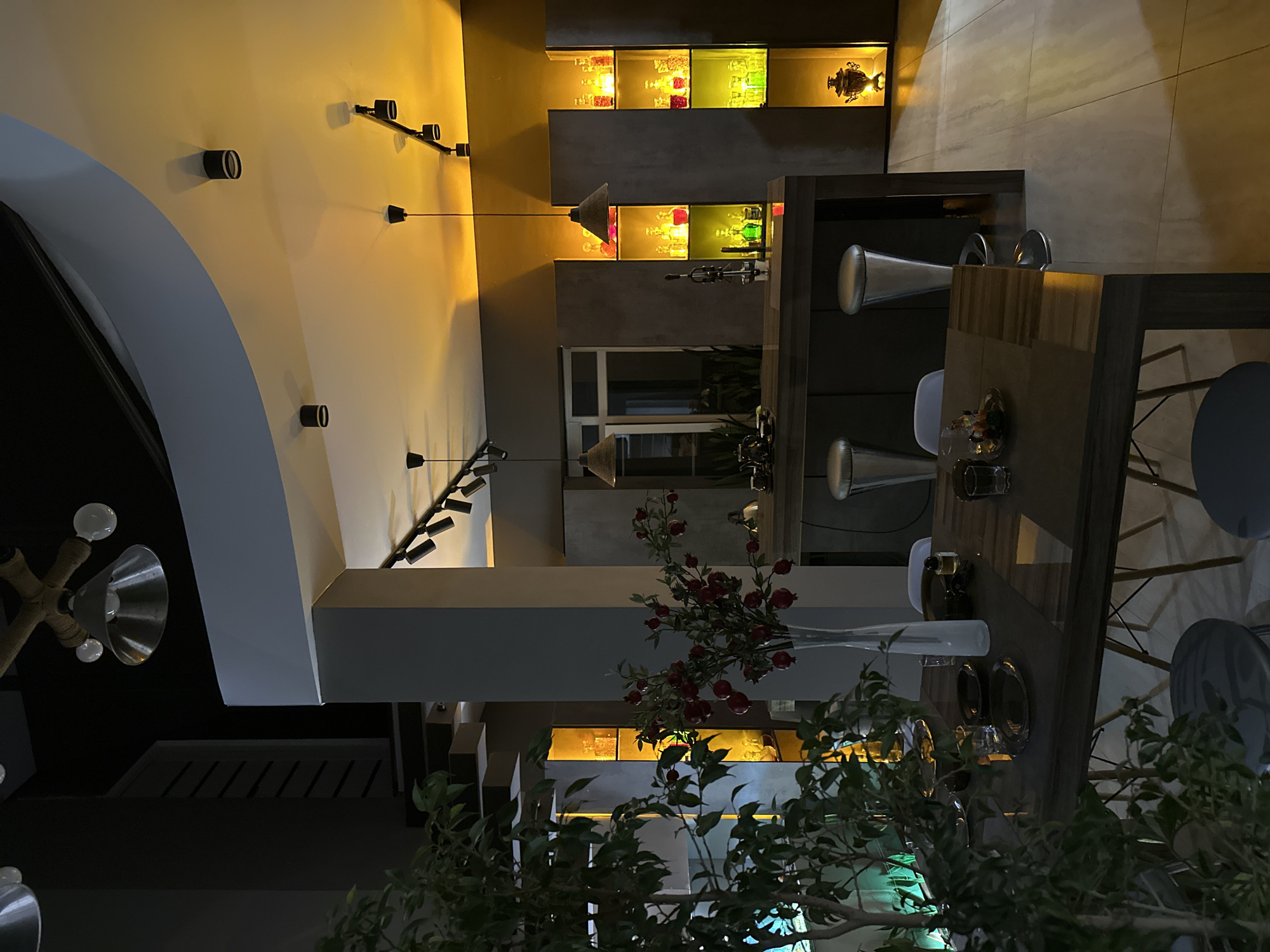The image depicts the interior of a restaurant or bar, likely located in Kursk, Russia. The ambiance is modern, with a focus on decorative lighting, including track lighting and pendant lamps. The scene includes a bar area with shelves displaying colorful bottles and glassware. In the foreground, there is a dining area with wooden tables set for meals. One table is adorned with a vase of red flowers and partially obscured by green foliage, suggesting an effort to create a natural aesthetic. The chairs are a contemporary design, and place settings are visible on the tables. The overall mood is dimly lit and suggests a relaxed or upscale atmosphere.