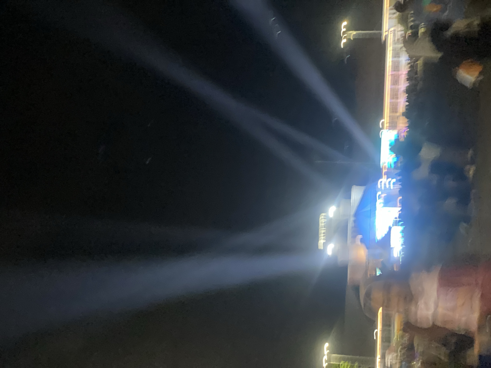 The image shows a nighttime outdoor event with stage lighting and an audience. Several bright spotlights emanate from the left side of the frame and fan upwards into the dark sky. In the midground, there is a stage or building structure with illuminated signage that appears to read "BALAI KOTA" and is lit by multiple bright lights. Below this structure, a large crowd of people is gathered, appearing to watch a performance or event. The foreground is occupied by more audience members, one of whom is partially visible on the left side of the image, appearing to be a child wearing a light-colored top. The overall scene suggests a public gathering or festival in an urban setting at night, likely in the city of Banjarbaru, Indonesia, given the text on the building.