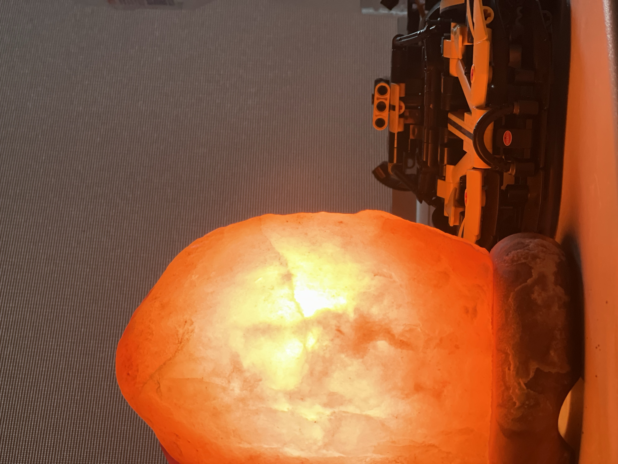The image displays an indoor scene featuring an illuminated Himalayan salt lamp alongside a detailed model. The dominant subject is a large, irregularly shaped Himalayan salt lamp, glowing with an intense orange and yellow light from within, highlighting its crystalline texture. Its warm radiance provides the primary illumination for the visible area.
To the right of the salt lamp, partially resting on a surface and partially obscured, is a complex mechanical model. This model appears to be an intricate Lego Technic set, specifically depicting the engine and chassis of a car, constructed from black and yellow/beige components. A notable detail on this model is a small, red circular sticker with the word "BUGATTI" clearly visible, identifying the brand of the modeled vehicle.
Adjacent to the main salt lamp, there is also a dark-colored, cylindrical object, possibly another salt lamp element or a decorative item, subtly lit by the warm glow. The background consists of a textured wall or surface, characterized by fine, vertical parallel lines, which could be wallpaper or paneling.
The lighting conditions suggest an evening or nighttime setting, with the salt lamp creating a cozy and warm ambiance. The image is oriented vertically, showing the objects positioned at the bottom and right, against the textured background on the left. No people are visible.