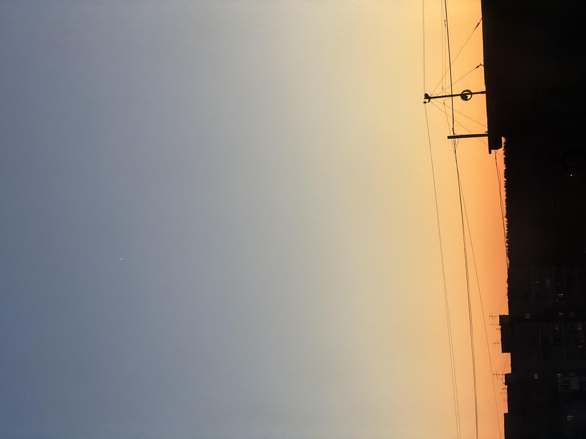 The image captures a sky gradient transitioning from blue to orange during what appears to be either sunrise or sunset. A utility pole with wires runs vertically along the right side, adjacent to a dark silhouette of buildings. There is a single, small bright dot, possibly a star or planet, visible in the bluer portion of the sky. The image is rotated 90 degrees counter-clockwise. The location is unspecified and information about Perm, Russia cannot be determined.