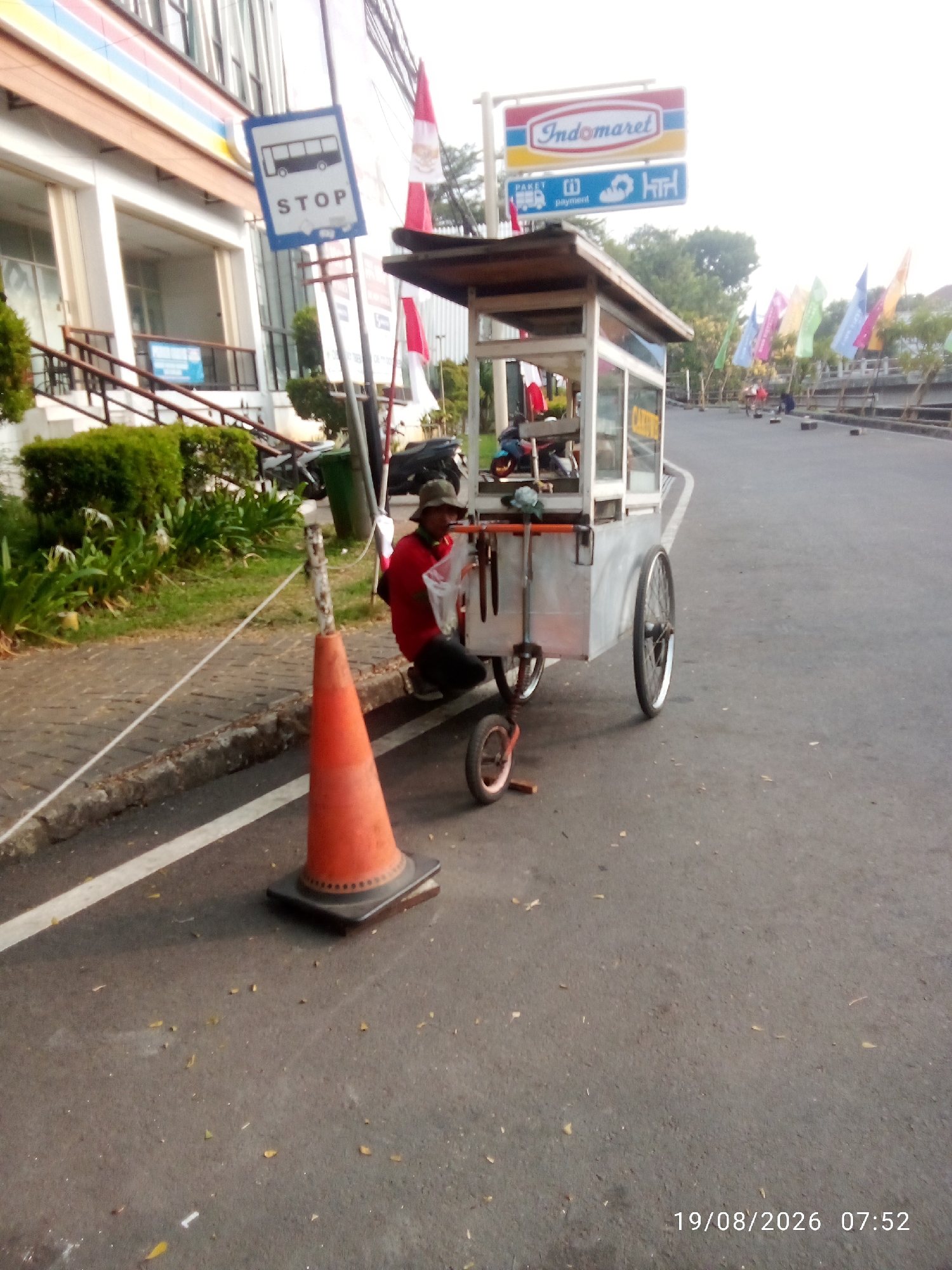 In East Jakarta, Indonesia, a man wearing a red shirt and a hat is seated on the sidewalk next to a food cart. The cart, made of aluminum, has a wooden roof and glass panels. A traffic cone is positioned on the road in front of the cart. In the background, a building with the Indomaret logo is visible, along with several flags. The scene appears to be captured on a street during daytime.