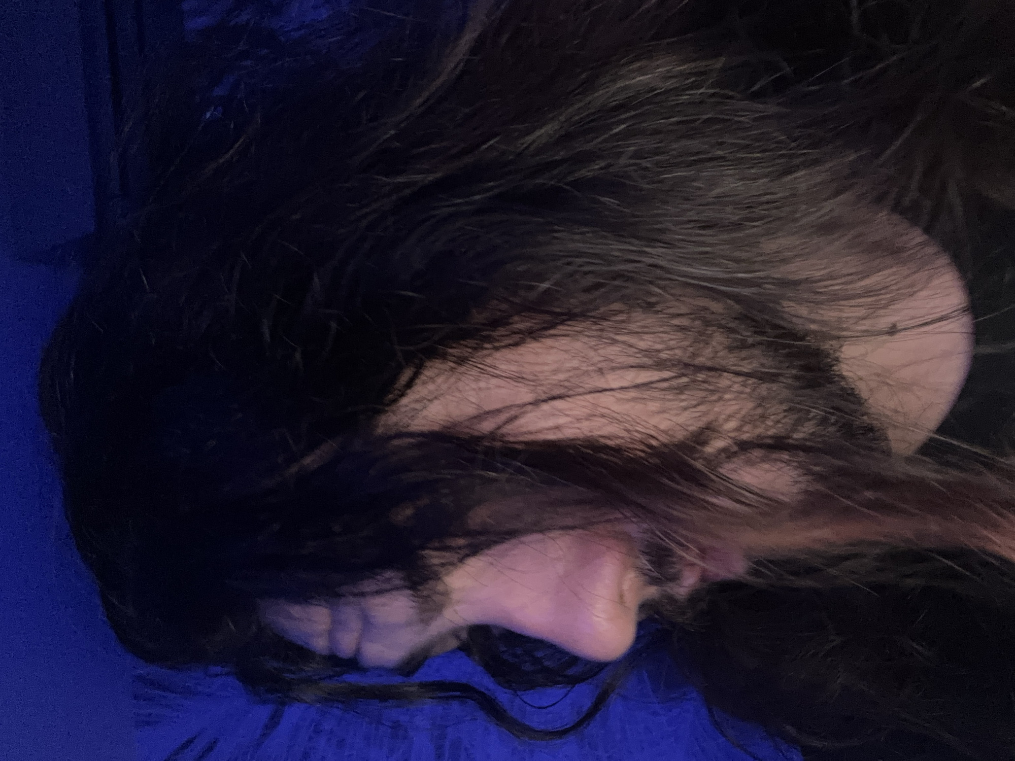 A close-up profile shot shows a person with long, dark hair that is partially obscuring their face. The person's head is tilted downwards and to the left. Their skin tone is fair, and they have stubble on their chin and jawline. The background is a dark blue, possibly indicating a dimly lit environment or a deliberate lighting choice. The image quality is somewhat grainy, and the focus is on the person's profile.
