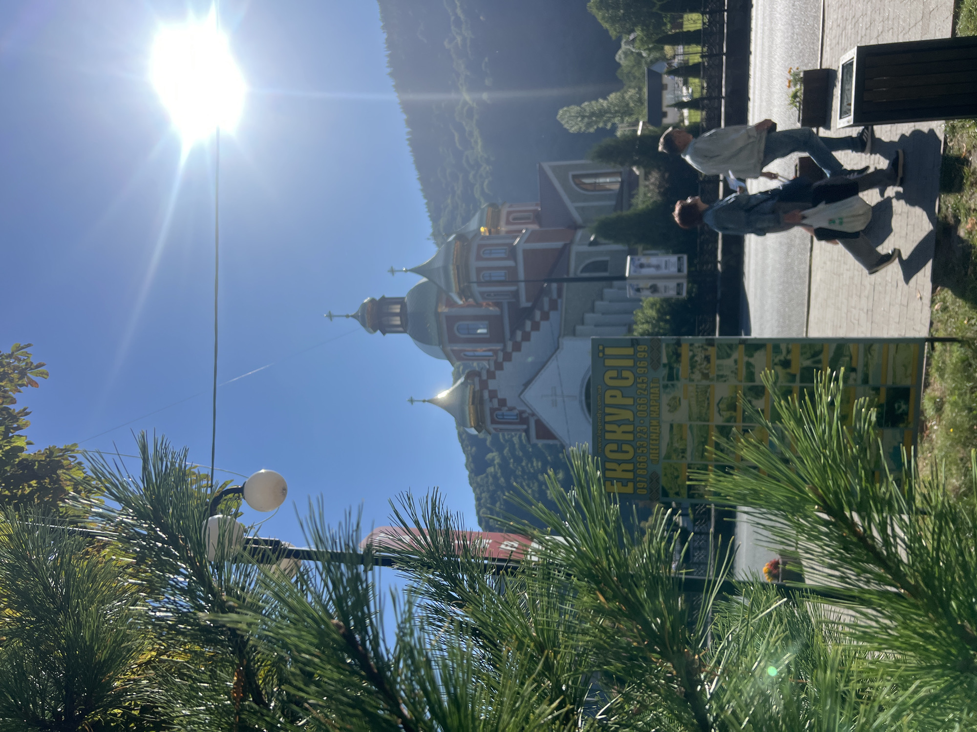 This is an outdoor, eye-level, portrait shot taken on a sunny day in Yaremche, Ukraine. In the foreground, a portion of a pine tree's branches is visible, obscuring the left side of the image. Behind the tree, a sign is prominently displayed, featuring the word "ЕКСКУРСІЇ" (Excursions) in large, yellow, bold lettering. Below this, a series of smaller images depict various tourist attractions, and phone numbers are listed. Two people are walking on a paved sidewalk to the right of the sign. A man in a light jacket and jeans is walking with his left leg forward, and a woman in a blue denim jacket and dark pants is walking beside him, carrying a white plastic bag in her left hand. They are both casting shadows on the sidewalk. Across the street, a large, ornate church with red and white walls and golden domes dominates the background. The church is situated on a hillside covered with lush green trees. The sun is shining brightly in the upper right portion of the image, creating lens flare. The sky is clear and blue.