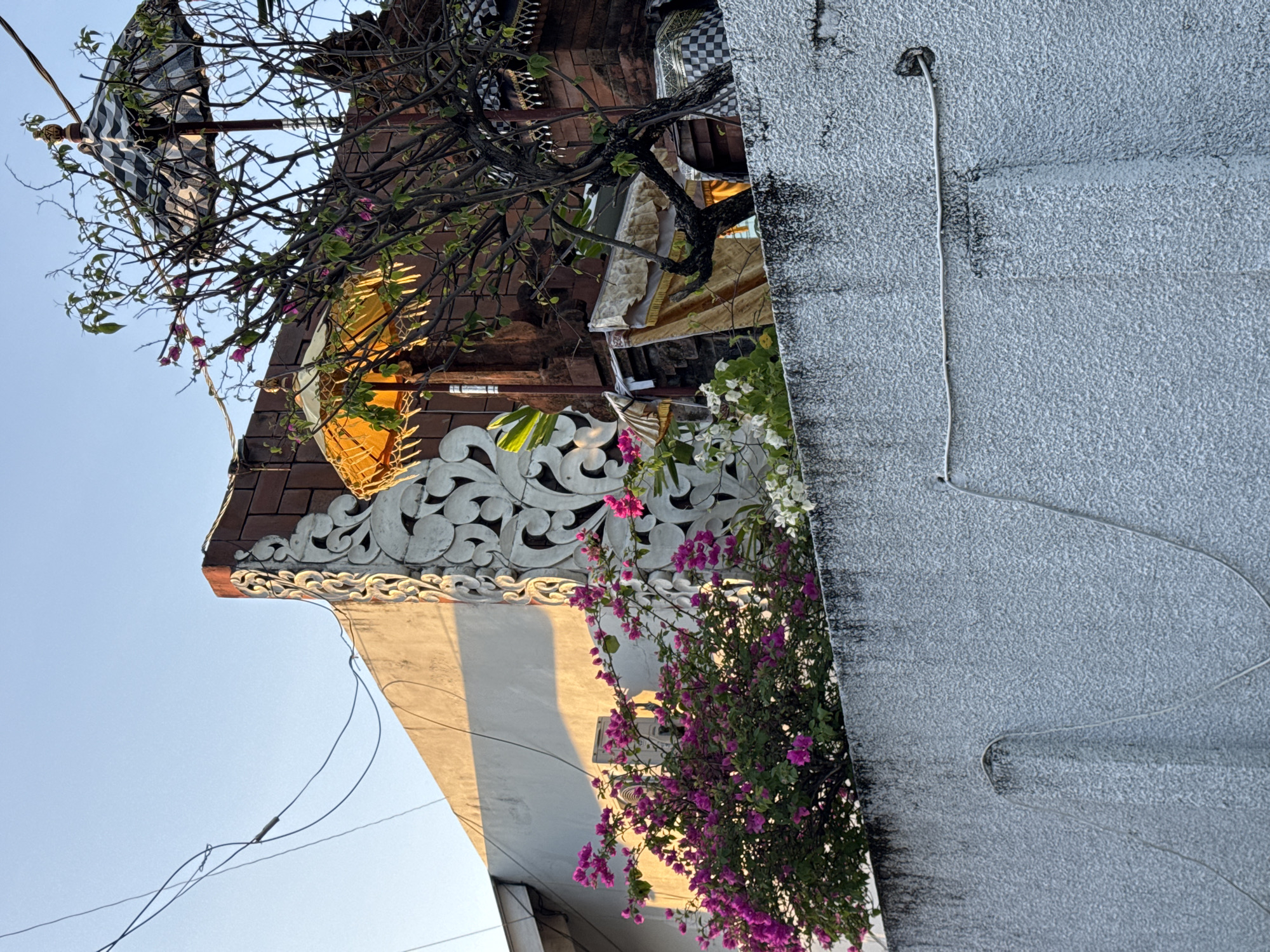 The image shows a partial view of buildings in Kuta, Indonesia, captured from a low angle. A white, textured wall dominates the right side of the frame, with a vertical wire running along its surface. To the left, a building with an ornate, white trim and a tiled roof is visible. A tree branch with foliage extends across the upper portion of the frame. Pink and white flowers cascade down the building's side. Above the ornate trim, a decorated umbrella is partially visible, suggesting a cultural or ceremonial significance. Electrical wires are visible against the clear sky.