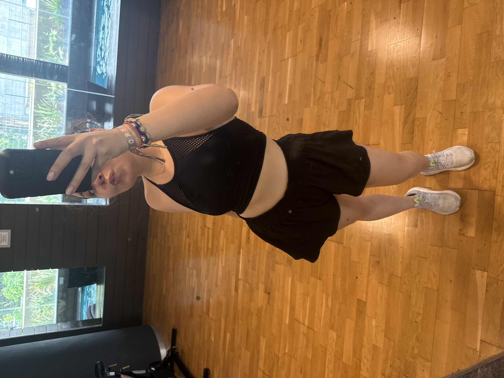 A person is taking a selfie in a room with wood floors, wearing a black sports bra, black shorts, and white athletic shoes. The room has large windows showing greenery and what appears to be a pool or water feature. In the background, a piece of exercise equipment is visible. The location is likely a gym or fitness studio. There's no explicit information or details suggesting that the location is in Mexico City, Mexico.