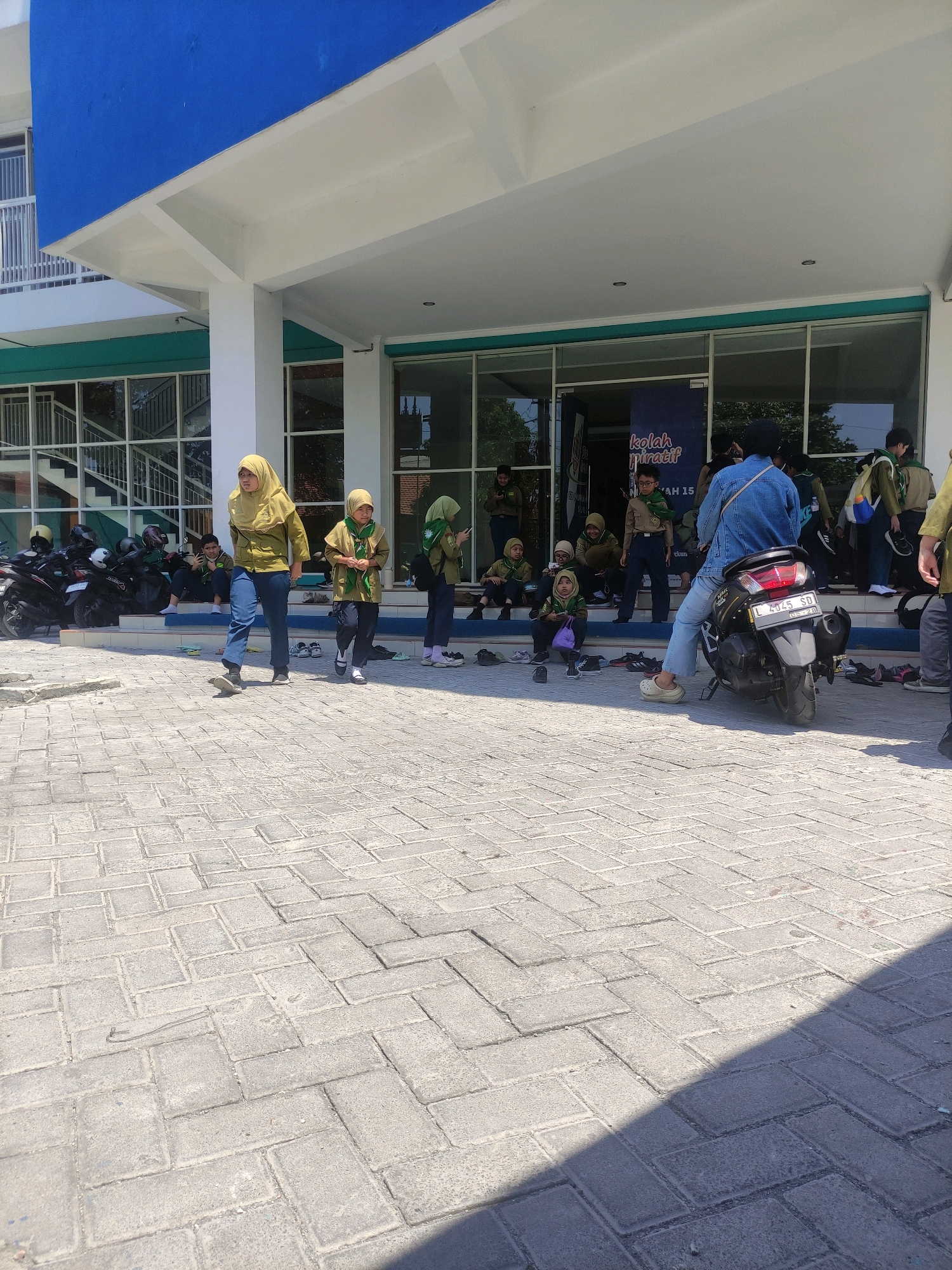 The image depicts an outdoor scene in front of a building with a blue and white facade. The foreground is a paved area with interlocking brick pavers, casting a shadow from an unseen object on the right. In the middle ground, a group of people, mostly young individuals wearing uniforms that appear to be scout attire with green scarves, are gathered. Some are sitting on steps leading up to an entrance, while others are standing or walking. One person in a denim jacket is seated on a black motorcycle parked on the paved area. A banner with Indonesian text, including "kolah miratif" and "AM 15," is visible through the glass doors of the building. Several motorcycles are parked to the left of the entrance. The overall atmosphere suggests a gathering or event, possibly at a school or educational institution, in Surabaya, Indonesia.