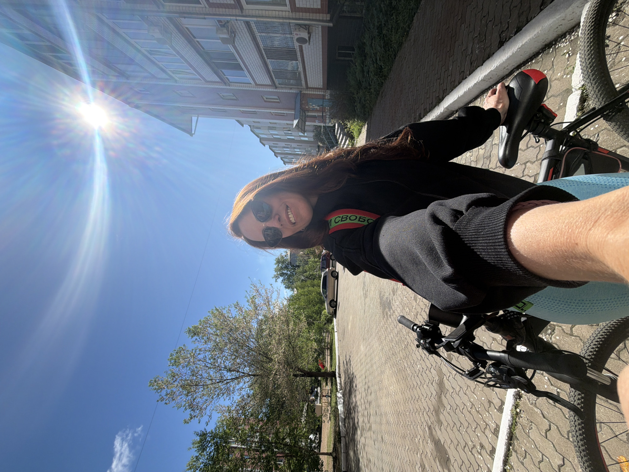 This diagonal selfie captures a person on a bicycle on a bright, sunny day in Birobidzhan, Russia. The main subject is a person, appearing female, with long, reddish-brown hair, wearing sunglasses and a black long-sleeved top, smiling cheerfully at the camera. They are positioned on a black bicycle, which features red accents on the seat and what appears to be a light blue or teal bottle holder or frame bag.
The setting is an urban or suburban street. To the left, a multi-story building with a brick facade, numerous windows, and some air conditioning units rises against the sky. The ground is paved with grey interlocking pavers, and a white line is painted along the edge of the path or road. Lush green trees, some exhibiting white blossoms, line the street, while other parked cars can be seen further down.
The time of day is clearly daytime, likely midday or early afternoon, given the brilliant sunshine and the sun's high position in the upper left corner, creating a strong starburst effect. The sky is a clear, vibrant blue with only a single wispy cloud visible.
A visible detail on the person's shoulder strap or bag is the partial text "СВОБО" in green letters on a red background. This is part of the Russian word "СВОБОДА," which translates to "FREEDOM." The image conveys a sense of enjoyment and freedom while cycling outdoors.
