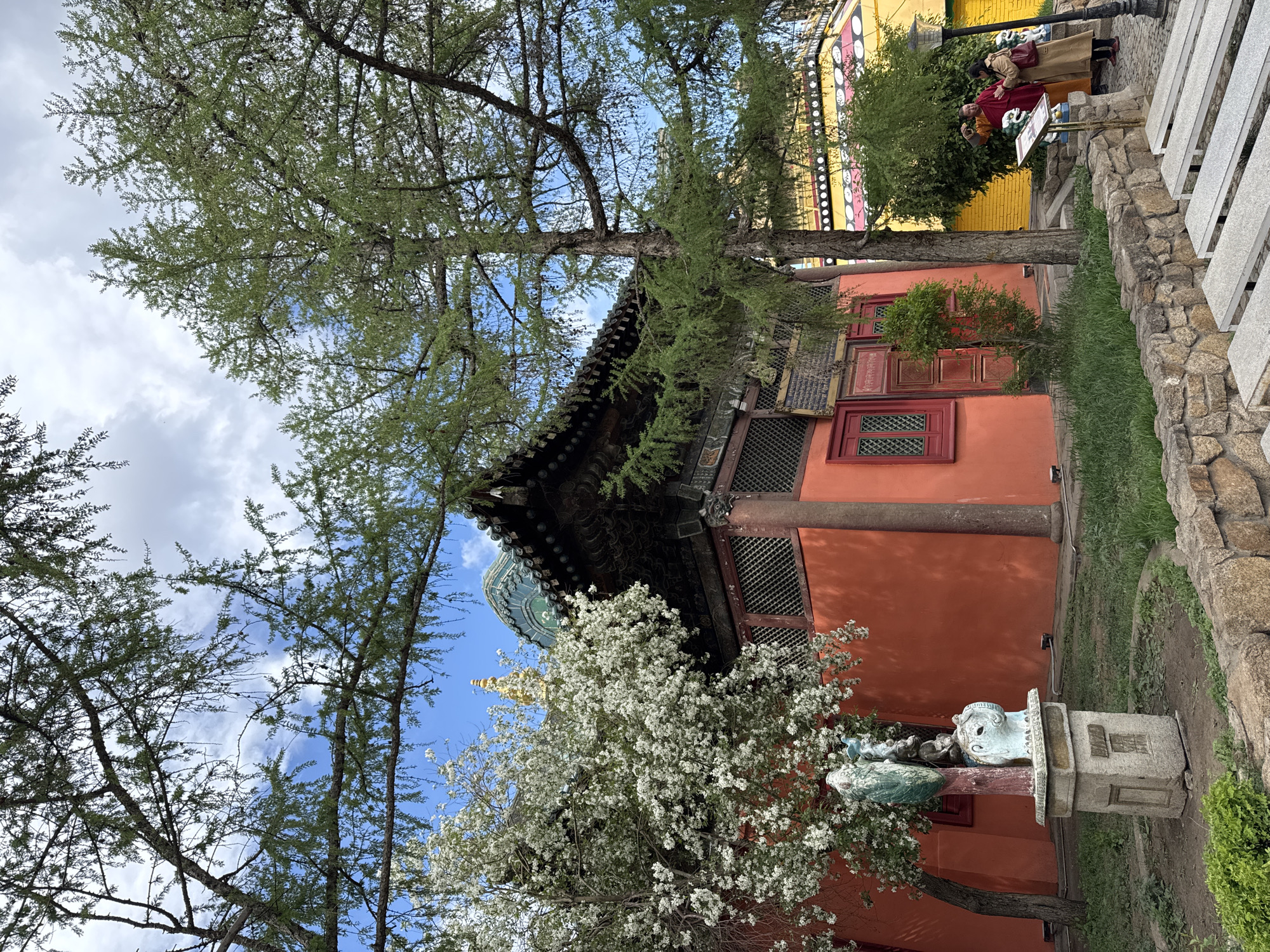 The image captures a scenic view of the Gandantegchinlen Monastery in Ulaanbaatar, Mongolia. An orange building featuring red-trimmed windows and a dark ornate roof is prominently displayed. A turquoise dome with gold accents peeks out from behind a large tree with white blossoms. To the right of the building, glimpses of a yellow structure and a couple of people can be seen. A stone sculpture on a pedestal stands in front of the building. The sky above is partly cloudy.