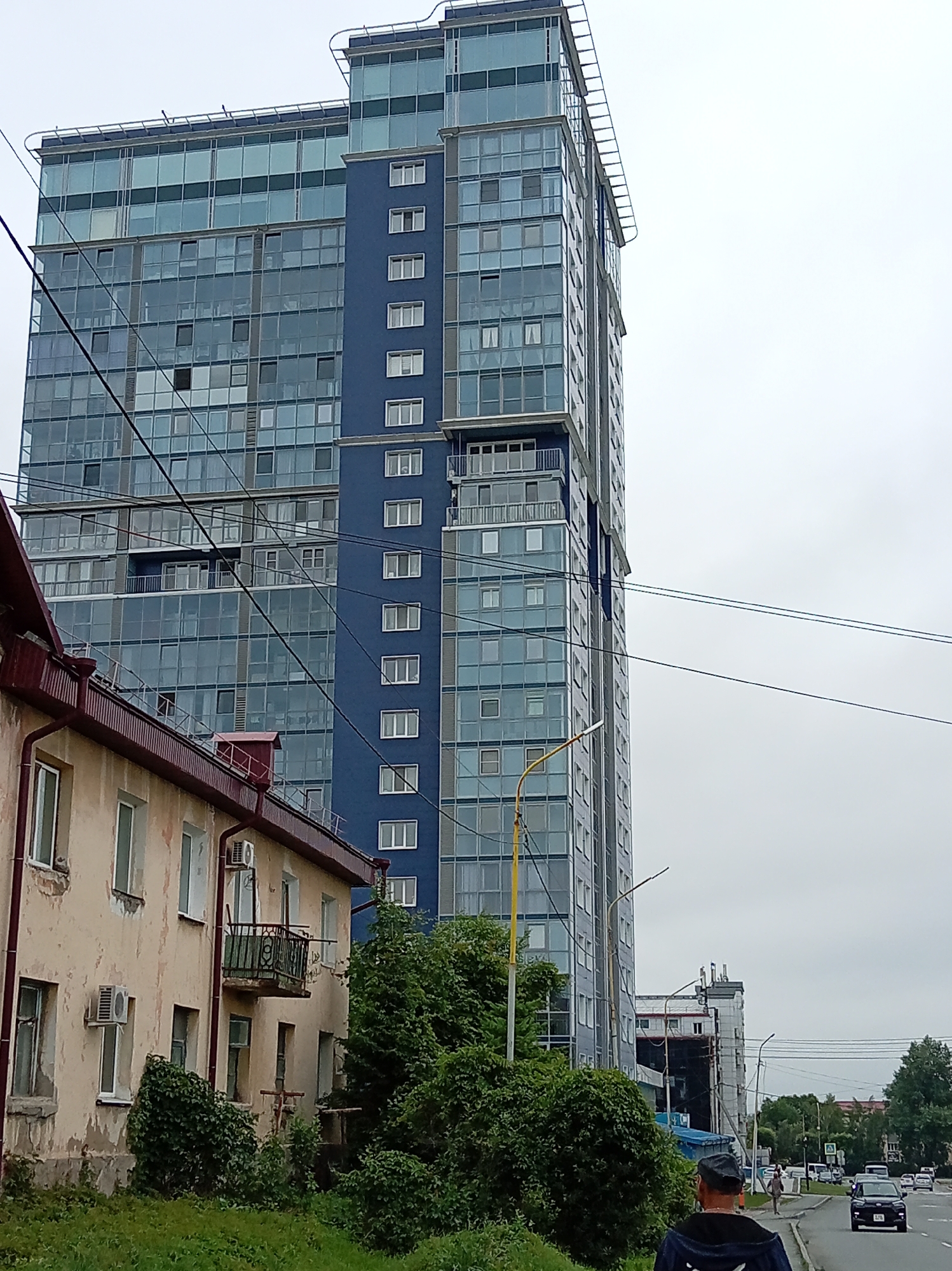 The image shows a street scene in Artyom, Russia, under an overcast sky. In the foreground, a weathered, light-colored apartment building with a dark red roof runs horizontally across the lower left of the frame. It has several windows and a small balcony with an ornate railing. Adjacent to this building and dominating the background is a tall, modern, blue and glass high-rise apartment building. Electrical wires are strung across the scene, obscuring parts of the buildings. To the right of the high-rise, a street with traffic is visible, with other buildings and trees further in the distance. A person wearing a dark hooded jacket and a cap is walking away from the camera on the sidewalk in the lower right corner.