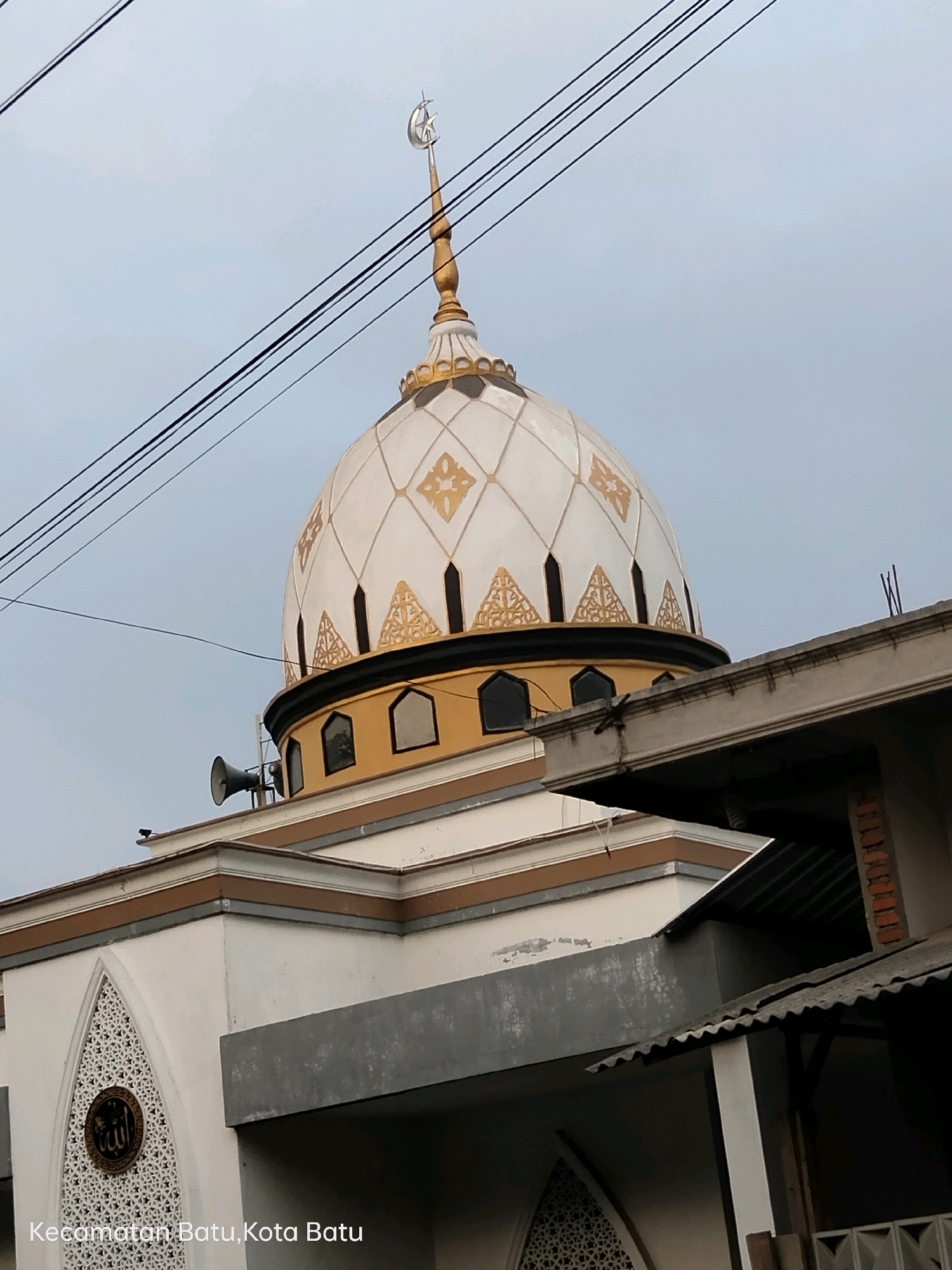 A close-up, slightly upward-angle shot showcases the white and gold dome of a mosque, topped with a crescent moon and star. Power lines crisscross the sky in the upper portion of the frame. The dome features decorative gold geometric patterns and small, dark windows. Below the dome, a yellow band encircles the mosque, interrupted by more dark windows. A loudspeaker is mounted on the left side of the dome's base. The lower part of the mosque's exterior is visible, with white walls, decorative arched elements, and a circular emblem with Arabic script. A grey roof structure partially obscures the right side of the building. The text "Kecamatan Batu, Kota Batu" is visible in the lower left corner, indicating the location in Batu City, Indonesia. The overall lighting suggests an overcast day. No people are visible in the image.