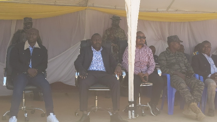 The scene is a gathering in Kirumba, Democratic Republic of Congo, held under a white tent with a yellow canopy. Six men are seated in a row. The man on the far left is wearing a black jacket, a blue shirt, blue jeans, and white sneakers. The man next to him is wearing a dark suit, a blue and white patterned shirt, black shoes. The man in the middle is wearing a red and white checked shirt, black pants and black shoes. The man on the far right is wearing a dark green camouflage uniform, brown boots, and a brown baseball cap. Two men are sitting behind the group, one is wearing camouflage military attire, the other is wearing civilian clothes. The environment is a dirt floor. The event is likely a formal gathering or a ceremony.