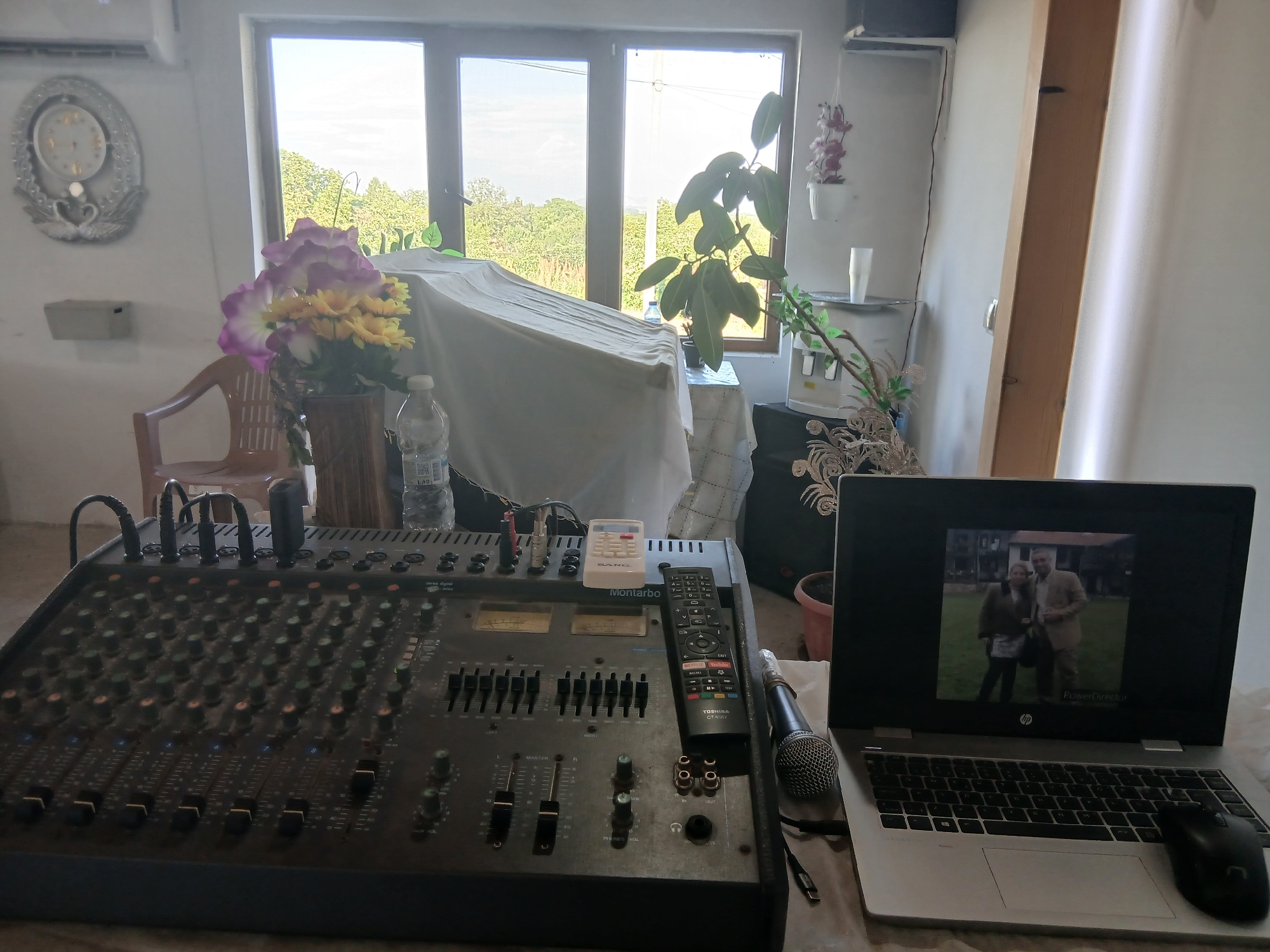 The image shows an indoor setting, likely a room used for audio mixing. Central to the scene is a large, black Montarbo audio mixer with numerous knobs, faders, and input/output jacks. A black Toshiba remote control rests atop the mixer near a wired microphone. Next to the mixer is an open HP laptop, its screen displaying a black and white photograph of a man and a woman. A black mouse is positioned to the right of the laptop.
In the background, a window provides a view of a landscape with trees, possibly indicating a location in the countryside. Part of the scene is obscured by a white tablecloth draped over an unseen object. To the left of the draped object, a flower arrangement in a vase sits on a surface along with a water bottle. A vintage-style clock hangs on the wall in the background on the left side of the window. A water dispenser, along with a potted plant is seen on the right side of the window.
Based on the details and context, the location is possibly a recording studio or an audio production setup in a residence, and given the latitude and longitude, the region is potentially Vetren Dol, Bulgaria.
