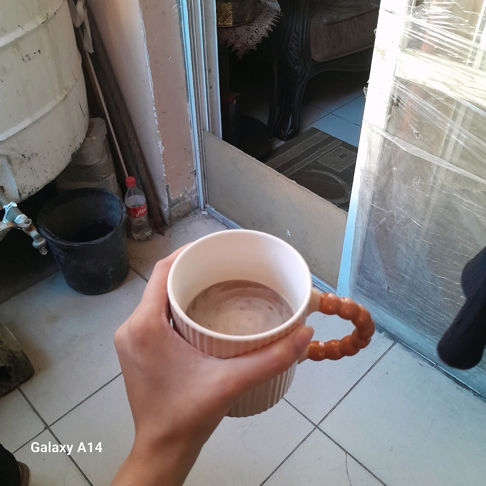 A close-up, eye-level shot captures a hand holding a ribbed, cream-colored mug filled with a light brown beverage, possibly coffee. The mug has two small, round, orange handles. The background is a mix of indoor and outdoor elements: a large white cylindrical object, possibly a water tank, sits on the left, adjacent to a black bucket and a partially consumed bottle of Coca-Cola. A faucet is visible on the side of the white object. The floor is tiled with light grey squares. Through an open doorway or window, an interior scene is partially visible, showing furniture covered with a lace cloth and another piece of furniture with dark, ornate carvings. The doorway itself is framed by a blue trim and covered with clear plastic sheeting. The lower left corner of the image displays "Galaxy A14" in white text, indicating the device used for capture. This scene is located in Khan Yunis, Palestinian Territories.