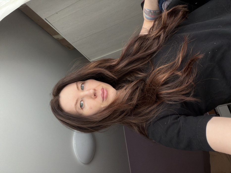A woman with long, wavy brown hair looks directly at the camera. Her eyes are light blue. She wears a black t-shirt. Part of a tattoo is visible on her upper arm. The background features a light gray wall with an oval light fixture. Above her is a white or light gray surface that appears to be a cabinet or shelf. The picture is rotated slightly counter-clockwise.