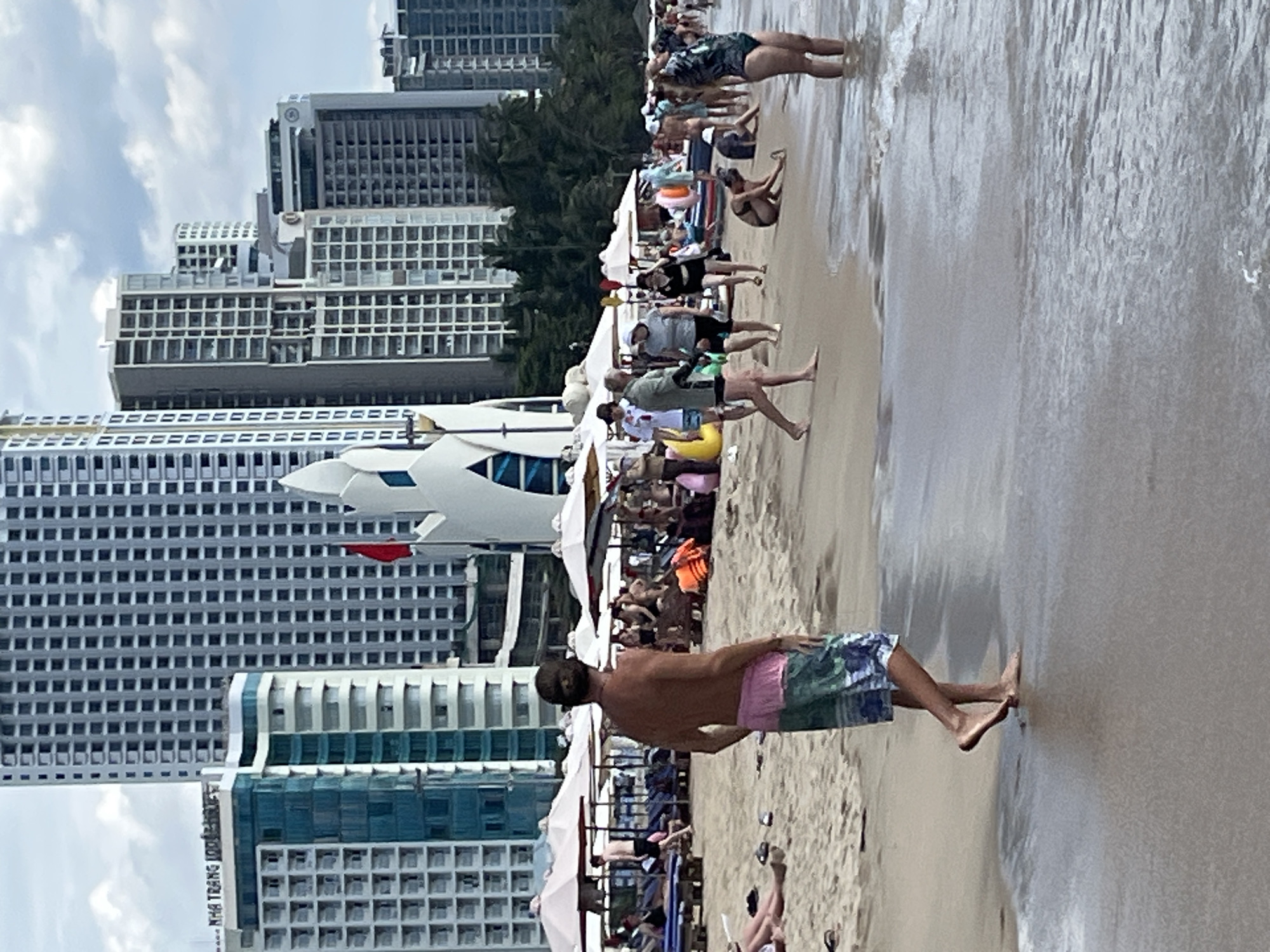 The image depicts a lively beach scene in Nha Trang, Vietnam, with the distinctive architecture of the city's hotels forming a backdrop. In the foreground, a man wearing colorful swim trunks stands facing away from the camera, with his back to the sea. The sandy beach is populated with numerous beachgoers enjoying the day. Several beach umbrellas and lounge chairs are set up, with some people relaxing under them. Others are walking along the shoreline, wading in the shallow water, or interacting with each other.
In the middle ground, a white, abstract sculpture-like structure is visible, a unique architectural element against the tall, modern buildings behind it. These buildings are characteristic of Nha Trang's beachfront development, with many stories and rows of windows. The sky is partly cloudy, suggesting a pleasant day for beach activities. The overall atmosphere is one of leisure and tourism, with the natural beauty of the beach blending with the urban landscape.