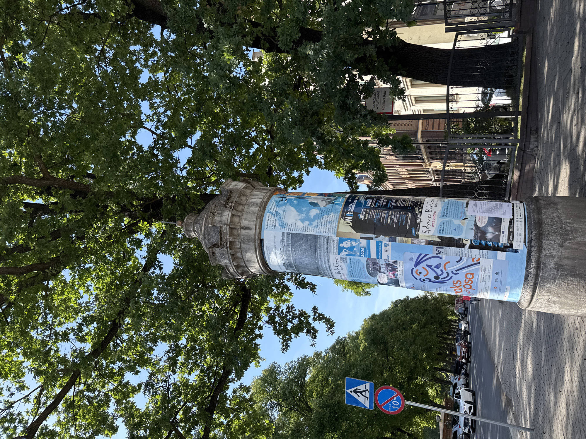 The image shows a street scene in Zgorzelec, Poland, on a sunny day. In the foreground, a tall, weathered concrete pillar is covered in various posters and flyers, advertising events and services. To the left of the pillar, a pedestrian crossing sign and a "10-minute parking" sign are visible. The street is lined with trees, casting shadows on the sidewalk. Cars are parked along the street and some are driving. In the background, a row of brick buildings can be seen. The scene is bustling with activity, suggesting a public urban space.