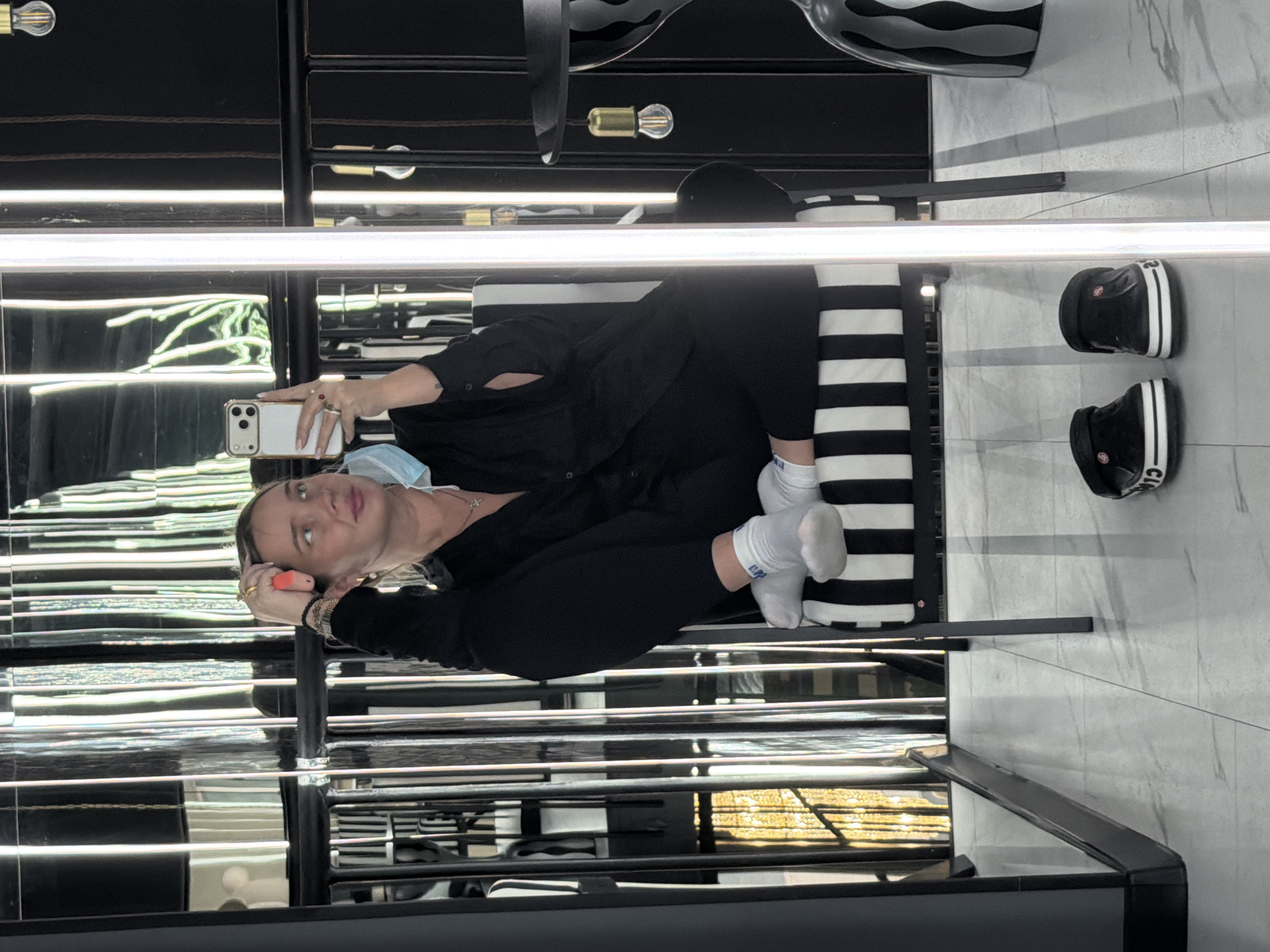 A person is taking a selfie while seated on a black and white striped bench. They are wearing black clothing and white socks. The person holds a white smartphone in their right hand, with a surgical mask partially obscuring their face. The environment features a mirrored or reflective wall and ceiling, with linear light fixtures illuminating the scene. Two black and white slides are positioned on the floor near the bench. The location is a commercial interior with marble flooring. The city and country are not discernible from the image.