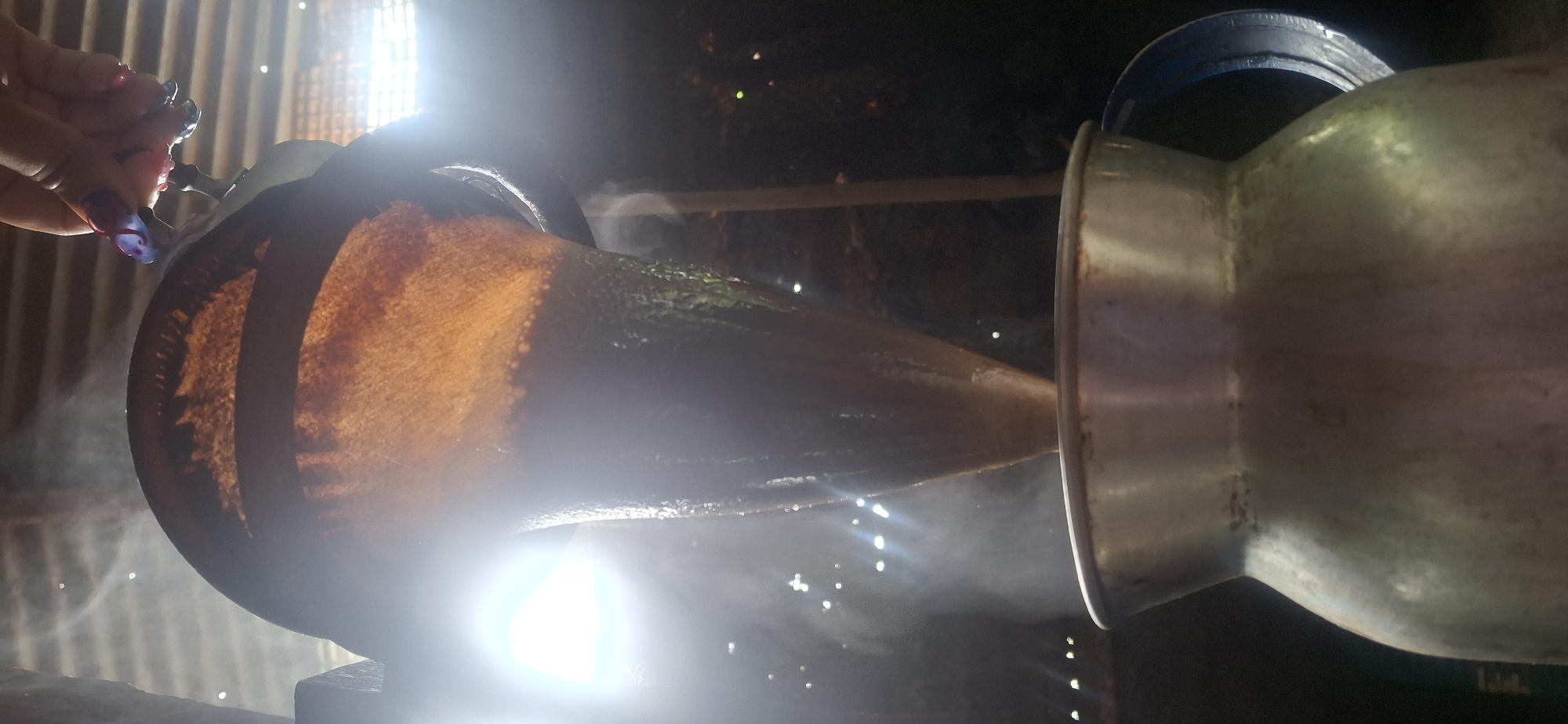 The image depicts a rustic scene, possibly related to traditional practices or cooking. A large, dark-colored vessel, possibly a pot or container, is connected to a smaller, metallic pitcher or pot. Steam or vapor is rising from the dark vessel, suggesting it contains something hot or is undergoing a process involving heat. A hand with painted fingernails is visible near the top left of the image, seemingly interacting with the larger vessel. The overall environment appears dimly lit and somewhat smoky.
The specific location, Vallecito, Venezuela, cannot be determined from the visual information in the image.