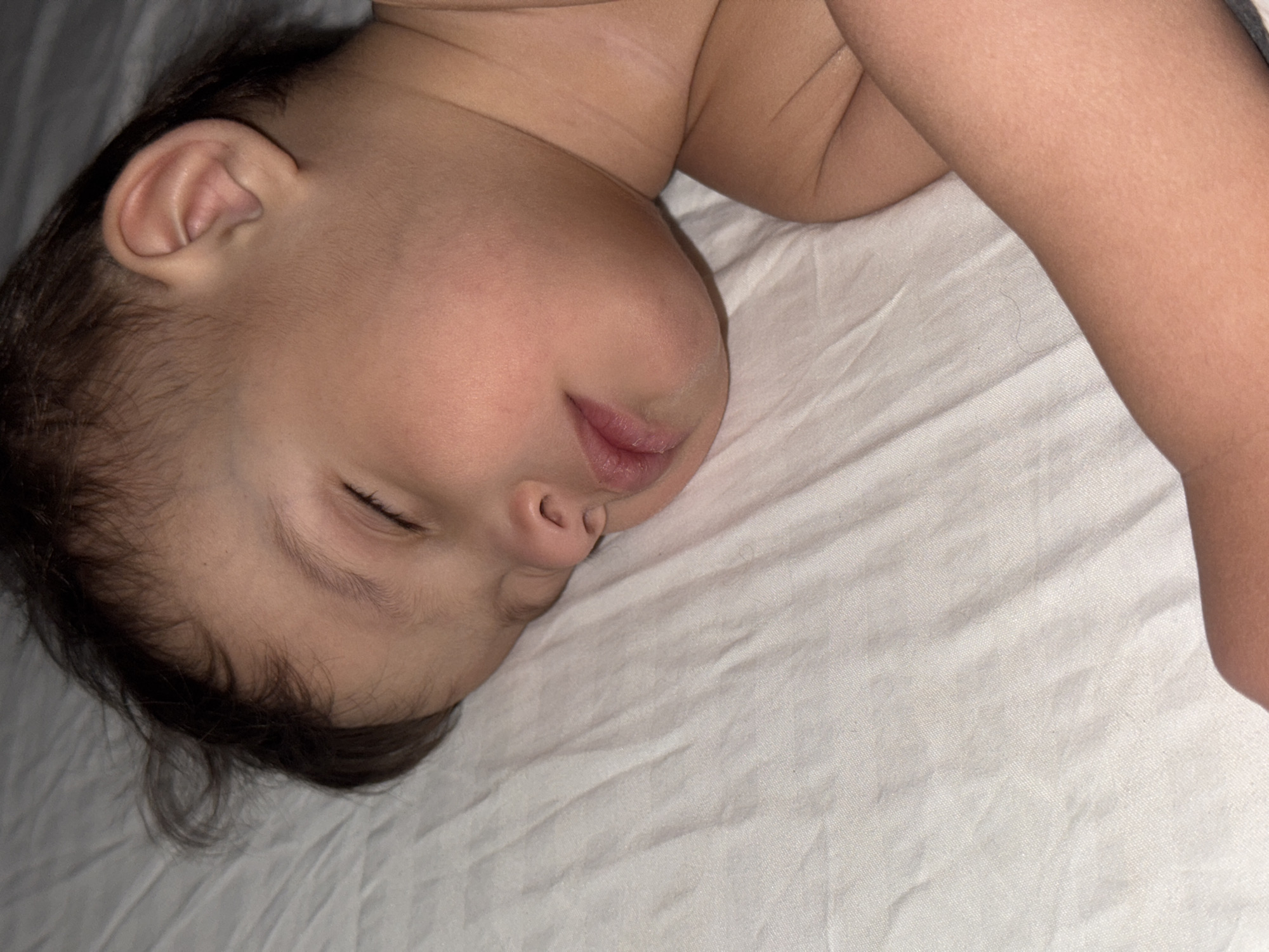 A baby is sleeping on a white sheet. The baby is lying on their side, with their face facing left. The baby's left arm is visible, resting on the sheet. The baby's hair is dark and short. The scene is indoors. There is no indication of the location being in the city of Chaikovsky, Russia.