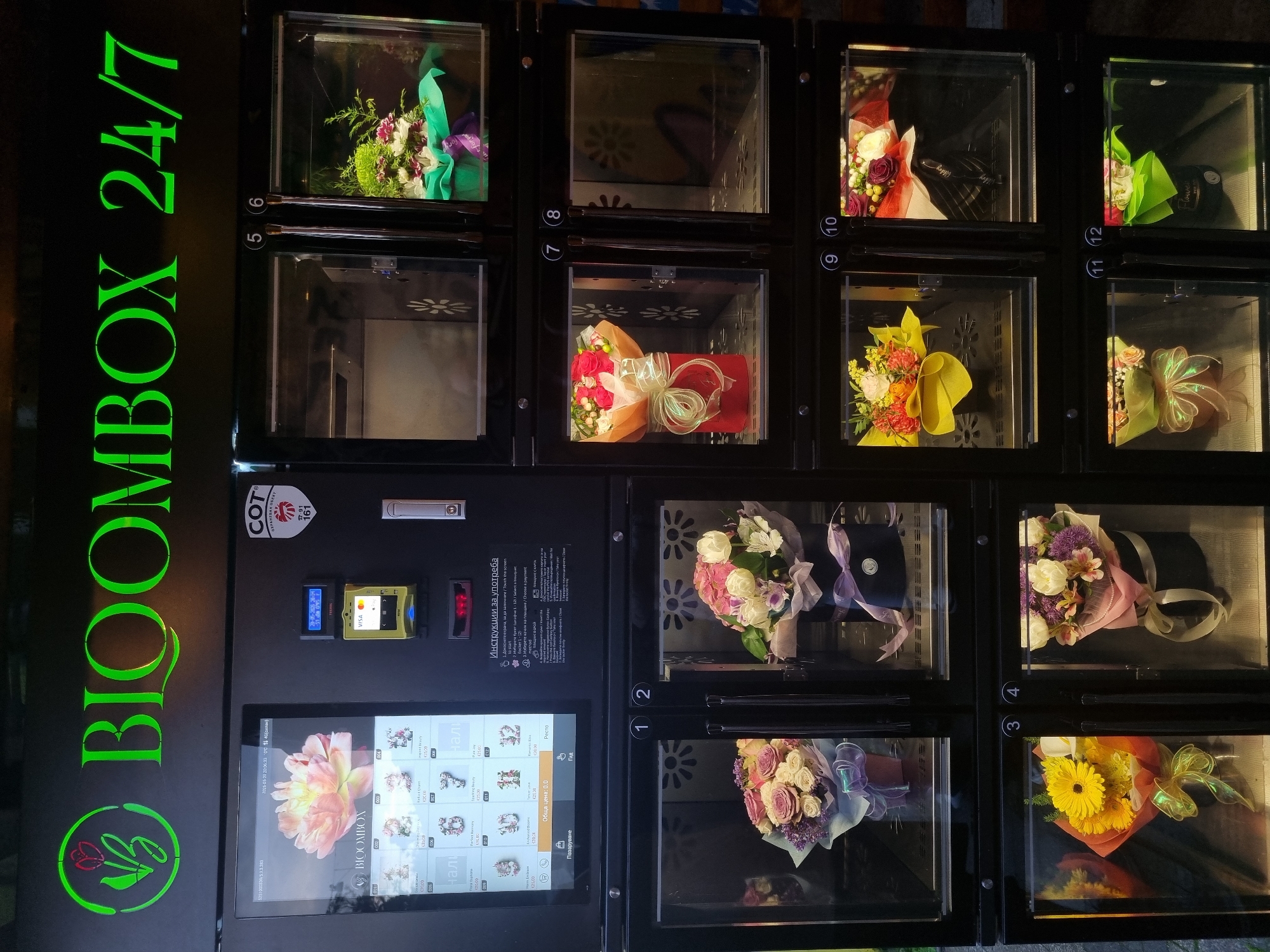 The image shows a BLOOMBOX 24/7 flower vending machine. The location is likely in Sofia, Bulgaria, due to the Cyrillic text on the screen. The machine has a large touch screen displaying flower options. It is located next to a payment system that accepts both cash and card payments. The vending machine has 12 compartments, each with a floral arrangement inside. The flowers are packaged and wrapped in cellophane. The machine displays the price of the flowers on the touch screen.