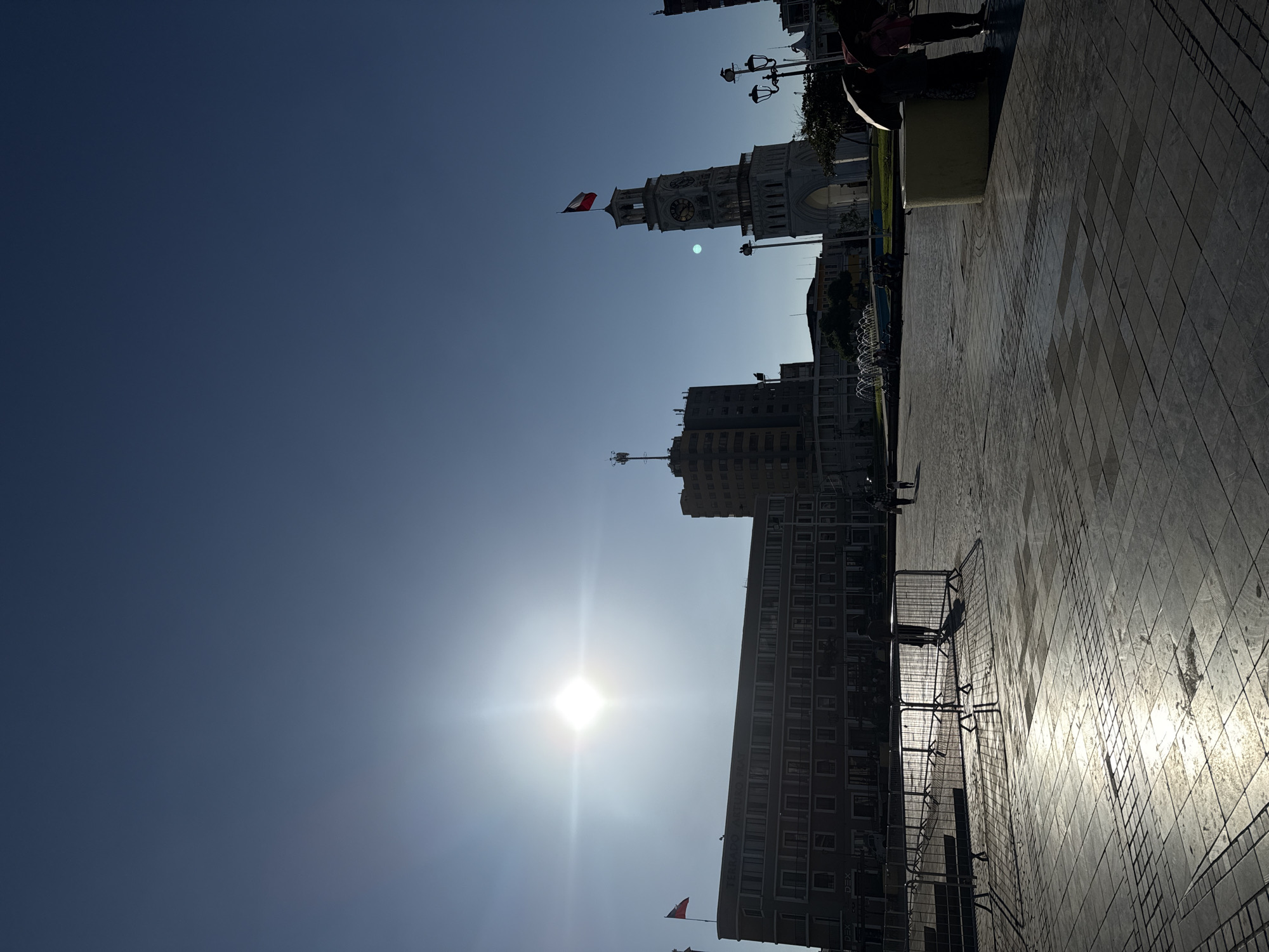 The image captures a bright, sunny day in Iquique, Chile, likely during the morning or late afternoon, given the low angle of the sun and the long shadows cast. The scene is set in a public plaza, paved with stone tiles that reflect the sunlight. In the background, a prominent clock tower stands tall, adorned with a Chilean flag. To the left of the tower, a modern building with multiple stories and balconies is visible, also flying a Chilean flag. Several other buildings of varying heights and architectural styles are present in the background.
In the foreground, temporary metal barriers are set up, possibly for an event or construction. A few people are scattered throughout the plaza. Two individuals are seen near the barriers, one appearing to be a security guard. Further in the distance, more people are walking, and a fountain can be observed. The sky is clear and blue, with the sun prominently featured and emitting a strong lens flare. The overall atmosphere suggests a busy urban environment on a clear day.