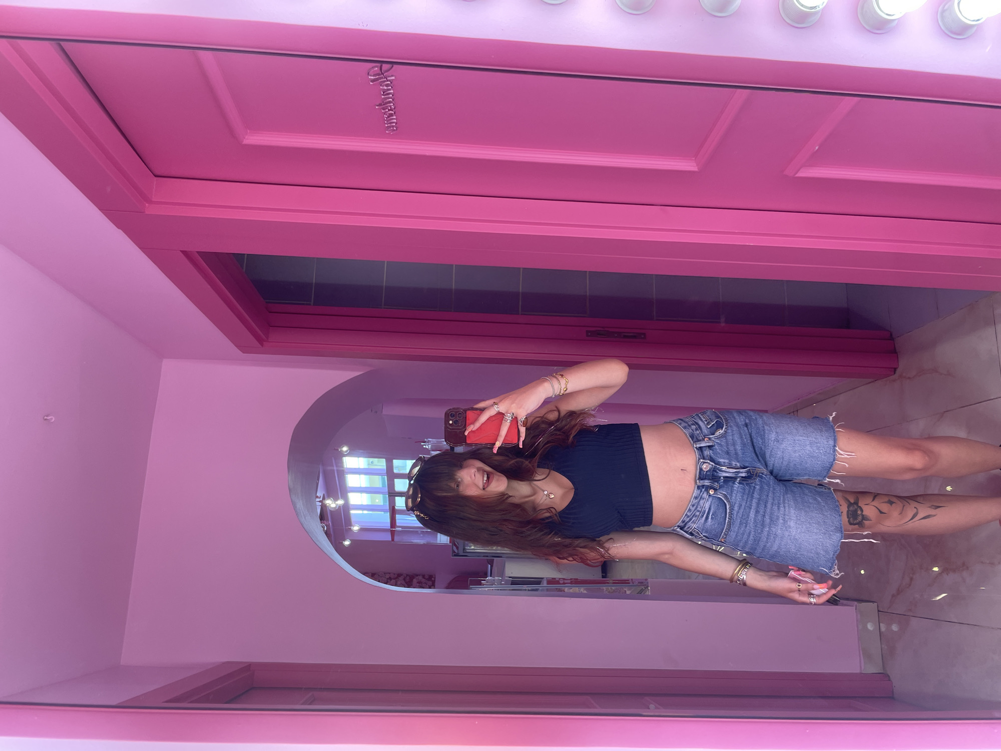 A young woman with long, wavy brown hair and bangs is taking a selfie in a mirrored room. She is wearing a dark, ribbed crop top and denim shorts with frayed edges. She has numerous rings on her fingers and bracelets on her wrists, and her nails are painted a bright pink. The room is decorated in various shades of pink, with a large mirrored wall that reflects the woman and the surroundings.
Behind her, the mirrored wall shows a glimpse of what appears to be a vanity area with multiple light bulbs lining the top of a pink vanity. The woman is holding a red smartphone in her right hand, taking the picture. Her left arm is bent at the elbow, and her hand is resting on her hip. She has a tattoo of a flower on her left thigh.
The setting appears to be an indoor space, possibly a dressing room or a boutique, given the vanity lights and the overall aesthetic. The lighting is bright, suggesting it could be daytime, or the room is well-lit by artificial lights. The mood conveyed by the woman's smile and her pose is cheerful and confident. The room's dominant pink color scheme creates a feminine and playful atmosphere. Visible text on the pink door in the background reads "Vogue Paris". The overall impression is that of a stylish and photogenic space.