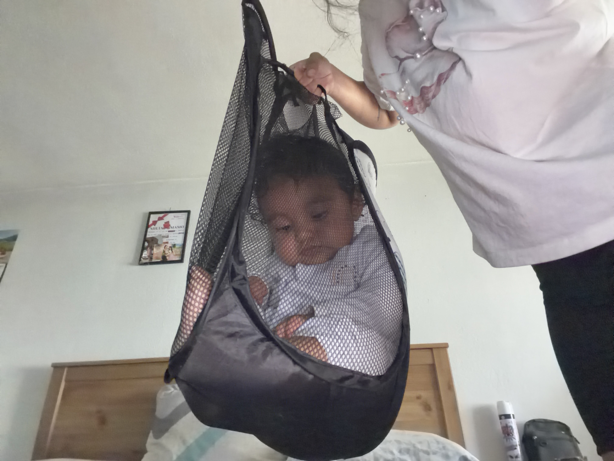 A baby is hanging in a black mesh laundry bag. The baby is looking down and appears to be calm. A person's hands are holding the top of the bag, supporting the baby. The baby is wearing a light blue long-sleeved shirt. The background shows a wall with a framed picture and a wooden headboard of a bed. The scene appears to be indoors, possibly a bedroom, with natural light coming from the right. There is no visible text in the image. The setting is likely a home in Razhevo Konare, Bulgaria.
