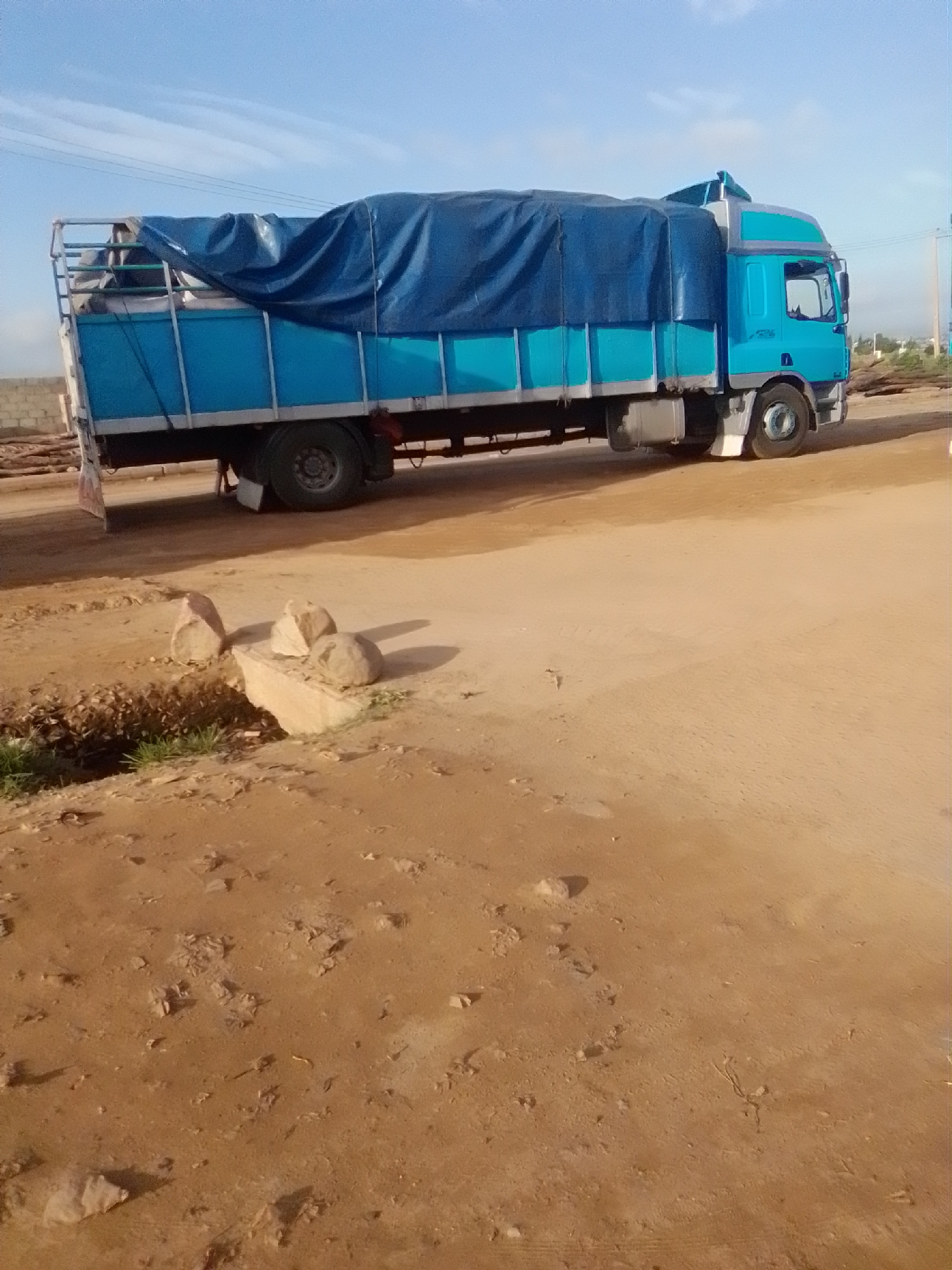An image shows a blue truck with a blue tarp covering its cargo. The truck is parked on a dirt road, and there are some rocks and debris on the ground. In the background, there is a brick wall and some trees. The sky is blue with some clouds. The scene appears to be in an outdoor setting. There are no people or animals visible in the image.