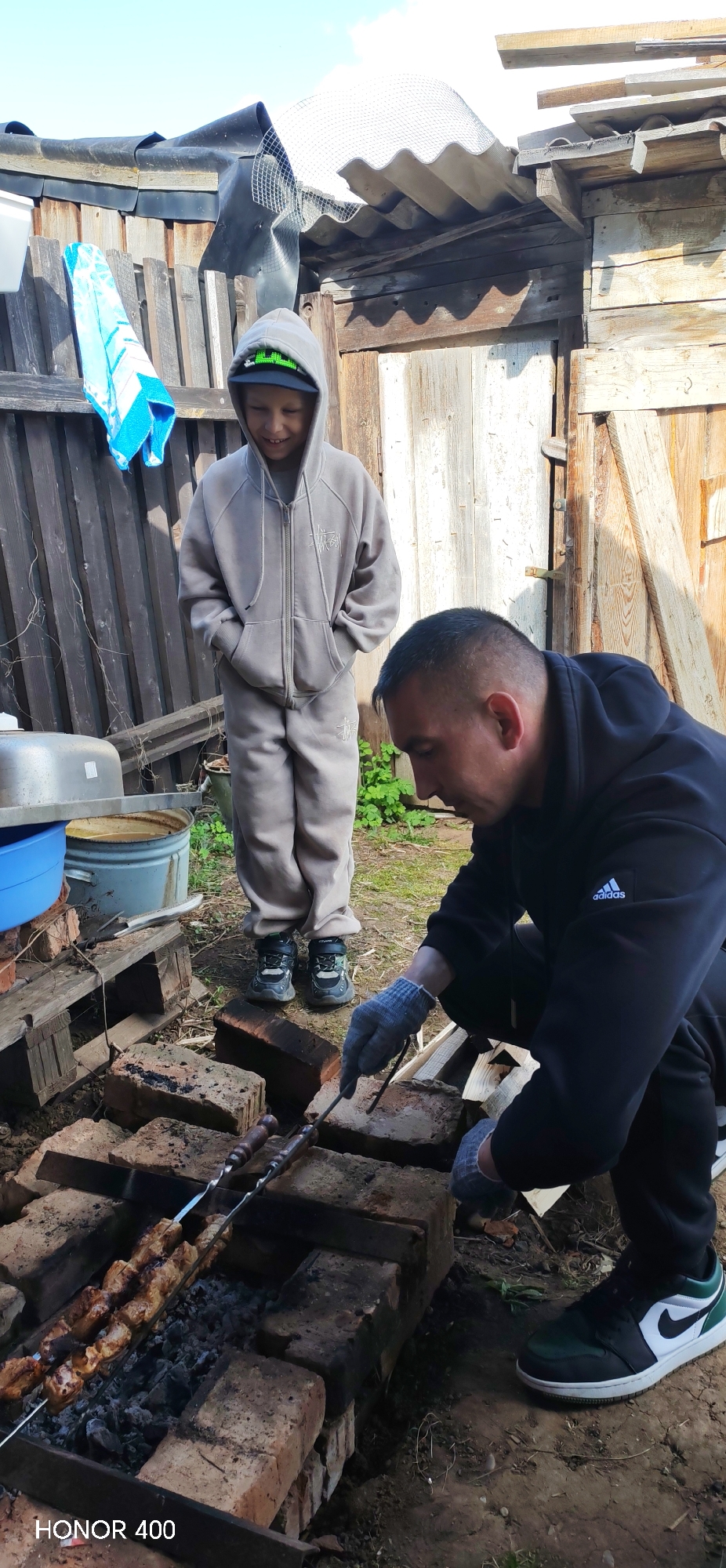 The image shows an adult male and a child in an outdoor setting, likely a backyard. The man, dressed in a black Adidas hoodie and dark pants, is crouched over a brick barbecue grilling kebabs on skewers. He is wearing gray gloves. The child, standing behind the man, is wearing a gray hooded tracksuit and a green baseball cap. The child is smiling. The environment is rustic, with a wooden fence and various containers and pallets visible in the background. The presence of the Honor 400 watermark at the bottom of the image suggests the photo was taken using that smartphone model. It is unclear whether the location is the city of Noviy, Russia, although the prompt suggests this is the case.