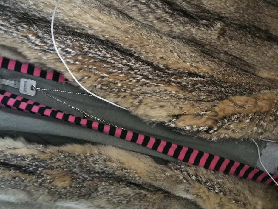 The image displays a close-up, angled view of layered garments and accessories. Dominating the frame is a multi-toned fur garment, presenting textured fibers in shades of light brown, dark brown, and black. Partially visible beneath or alongside the fur is a flat grey fabric, possibly an undergarment or lining. Vertically positioned within the frame, a strap or ribbon with alternating horizontal stripes of bright pink and black is evident. Draped over these layers are two metallic chains. One is a silver-colored bead chain, from which a silver-colored house key with a rectangular head and central circular hole is suspended. The second is a finer, silver-colored link chain, partially obscured, with a small, indistinct charm. A thin, white cable crosses the upper left quadrant of the image. No individuals are visible, and the specific location within the city of Paris, France, cannot be determined from the visual content.