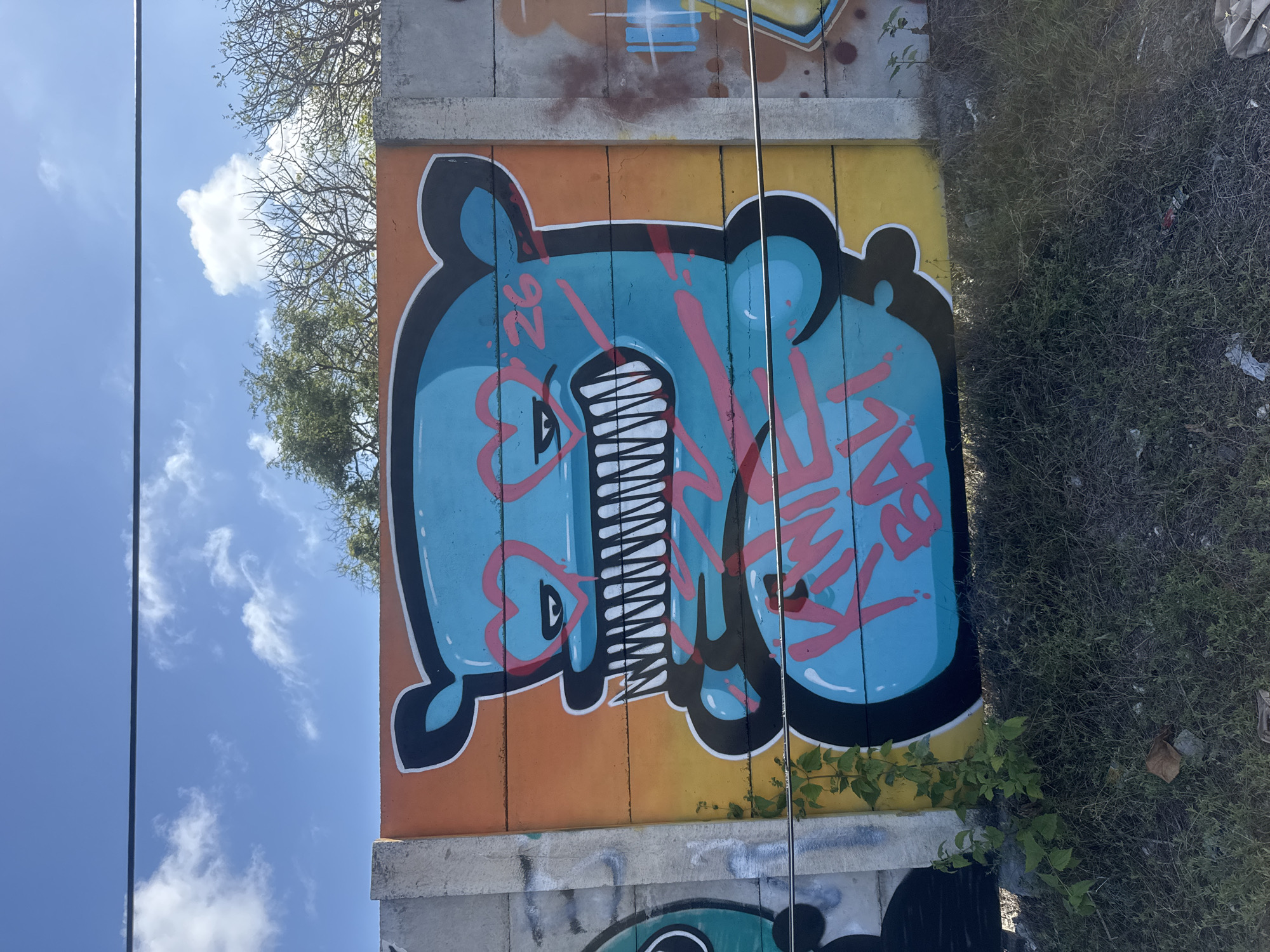 The image depicts a vibrant graffiti mural on a concrete wall under a bright blue sky with scattered white clouds. The mural features a large, stylized blue character with sharp teeth, heart-shaped eyes outlined in pink, and pink lettering that appears to read "KING BALL" across its torso. The character is set against a background gradient of orange and yellow. To the left of the main mural, a portion of another graffiti piece in teal and black is visible, and to the right, remnants of other graffiti in brown and blue are present. The wall itself is constructed from concrete blocks. Green vegetation, including small trees and ground cover, grows at the base of the wall. A thin black wire runs horizontally across the lower portion of the image, in front of the mural. The scene is an outdoor environment, likely in a public space, consistent with urban art installations. The location is identified as Kuta Selatan, Indonesia. There are no people or active interactions visible in the image.