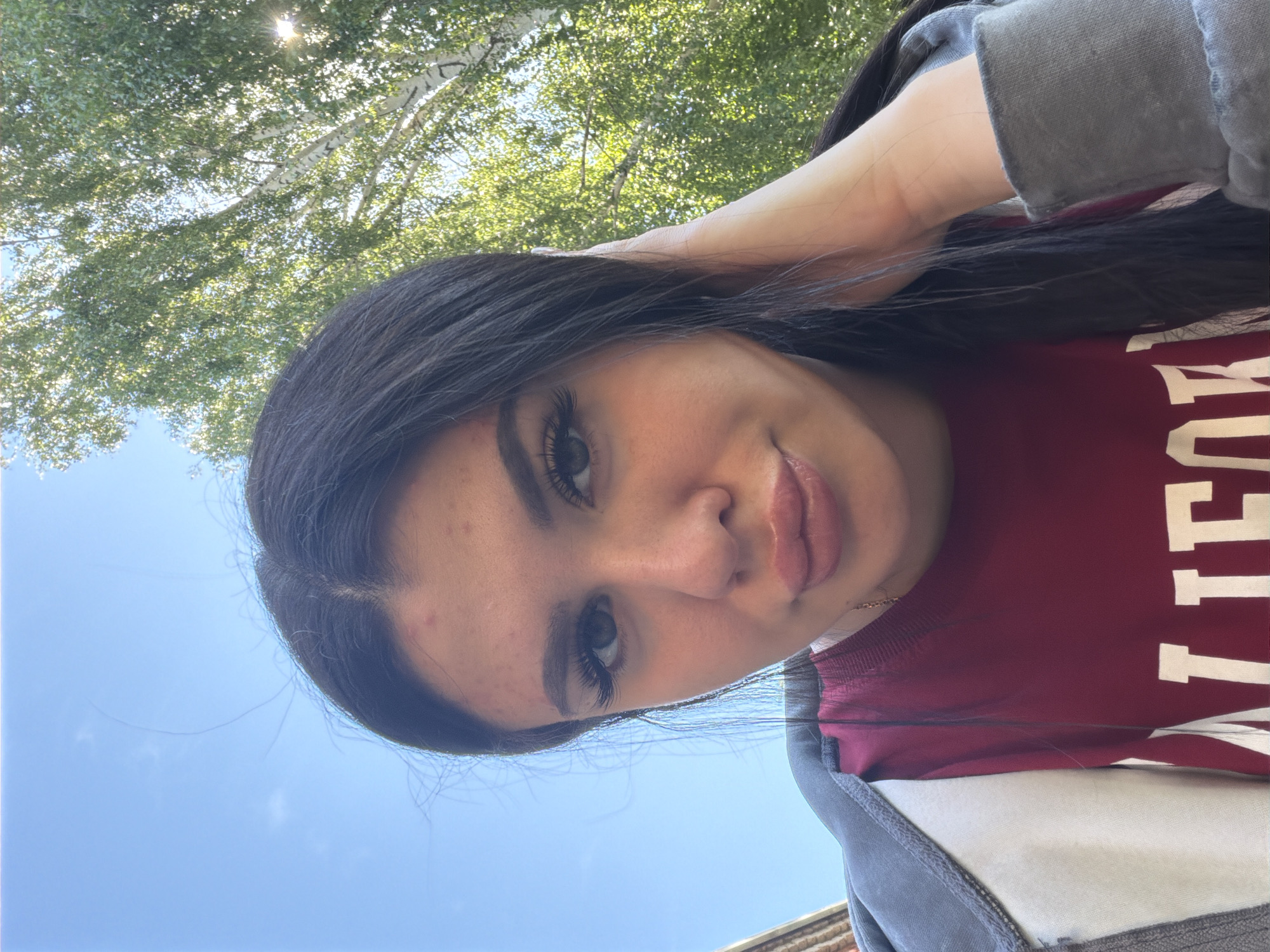 A young woman with long dark hair and dramatic eyelashes is captured in a selfie outdoors. She is wearing a maroon sweatshirt with white lettering that appears to spell out "UNIVERSITY". The background shows lush green trees with sunlight filtering through the leaves, and a clear blue sky. The woman's left hand is resting on her head, and she is looking towards the camera with a slight smile. The overall impression is casual and relaxed, possibly suggesting a warm, sunny day. There are no obvious location cues specific to Фоки, Russia, but the presence of trees and a bright sky indicate an outdoor setting during daylight hours.