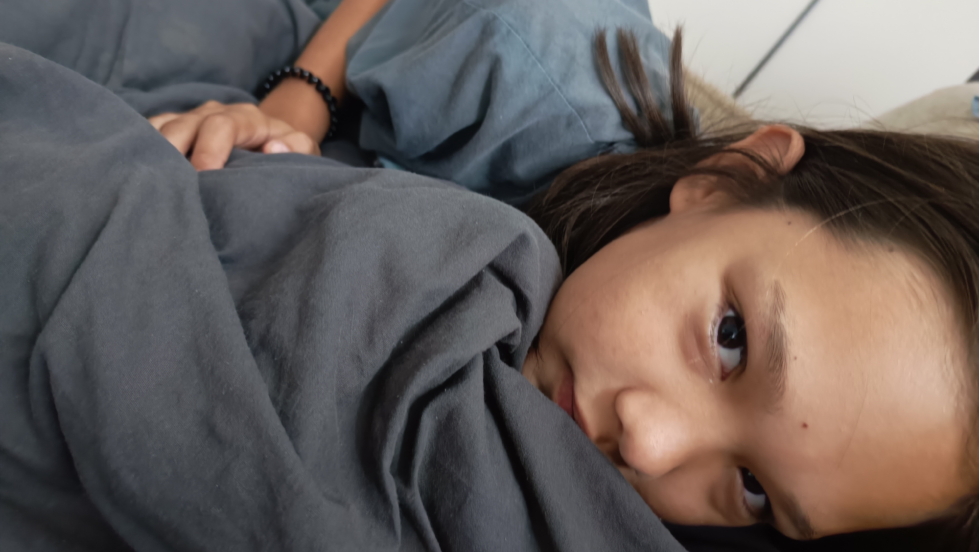 A person lies on a bed, partially covered by a dark grey blanket and a blue garment. The person's face is turned towards the camera, with their left eye visible. A black beaded bracelet is on their left wrist, which is resting on the blue garment. The background is out of focus and appears to be a room. There is no information in the image to determine a specific location in Karsha, Russia.