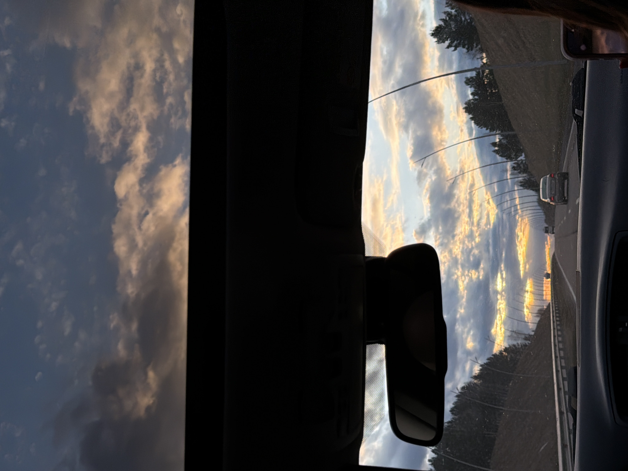Captured from within a vehicle on a highway, the image shows a view through the front windshield. The sky is filled with clouds illuminated by the setting sun, casting golden hues. The road stretches ahead, lined with trees and lampposts. Other vehicles can be seen in the distance. A rearview mirror is visible within the car. Part of a cell phone and the silhouette of a person's head are visible on the right side. The location is likely near Yemelyanovo, Russia, given the context.