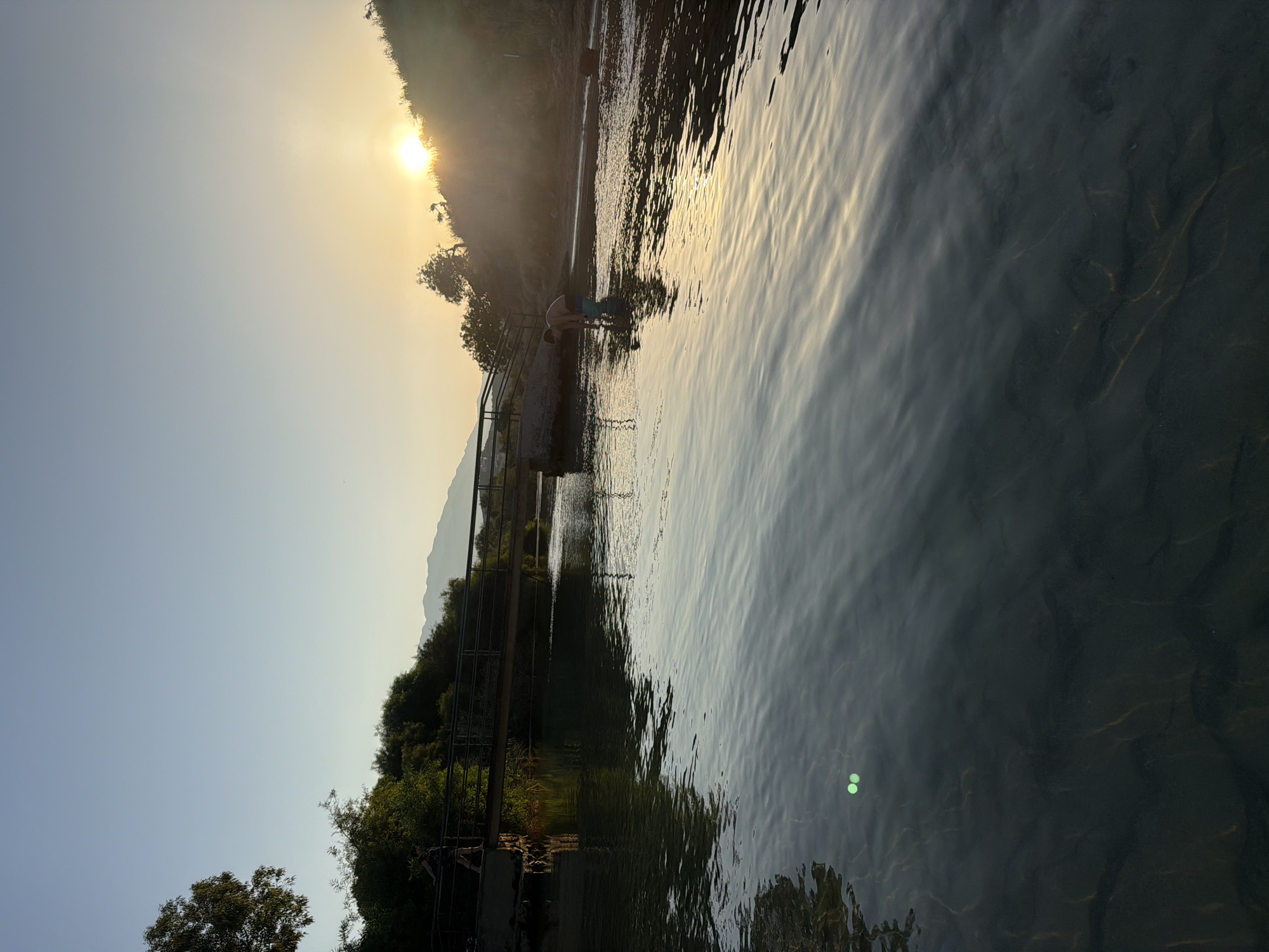 A serene body of water reflects the golden hues of a sunset as a person stands in the shallow water near the riverbank. The sun, low on the horizon, casts a warm glow over the scene, illuminating a nearby bridge arching over the water. Lush greenery lines the opposite bank, with distant mountains forming a hazy backdrop. The water itself is clear, revealing the sandy bottom beneath. Two small, bright green objects are visible submerged in the foreground of the water. This is the Almyros River in Georgioupoli, Greece, a popular spot for its natural beauty and tranquil atmosphere.