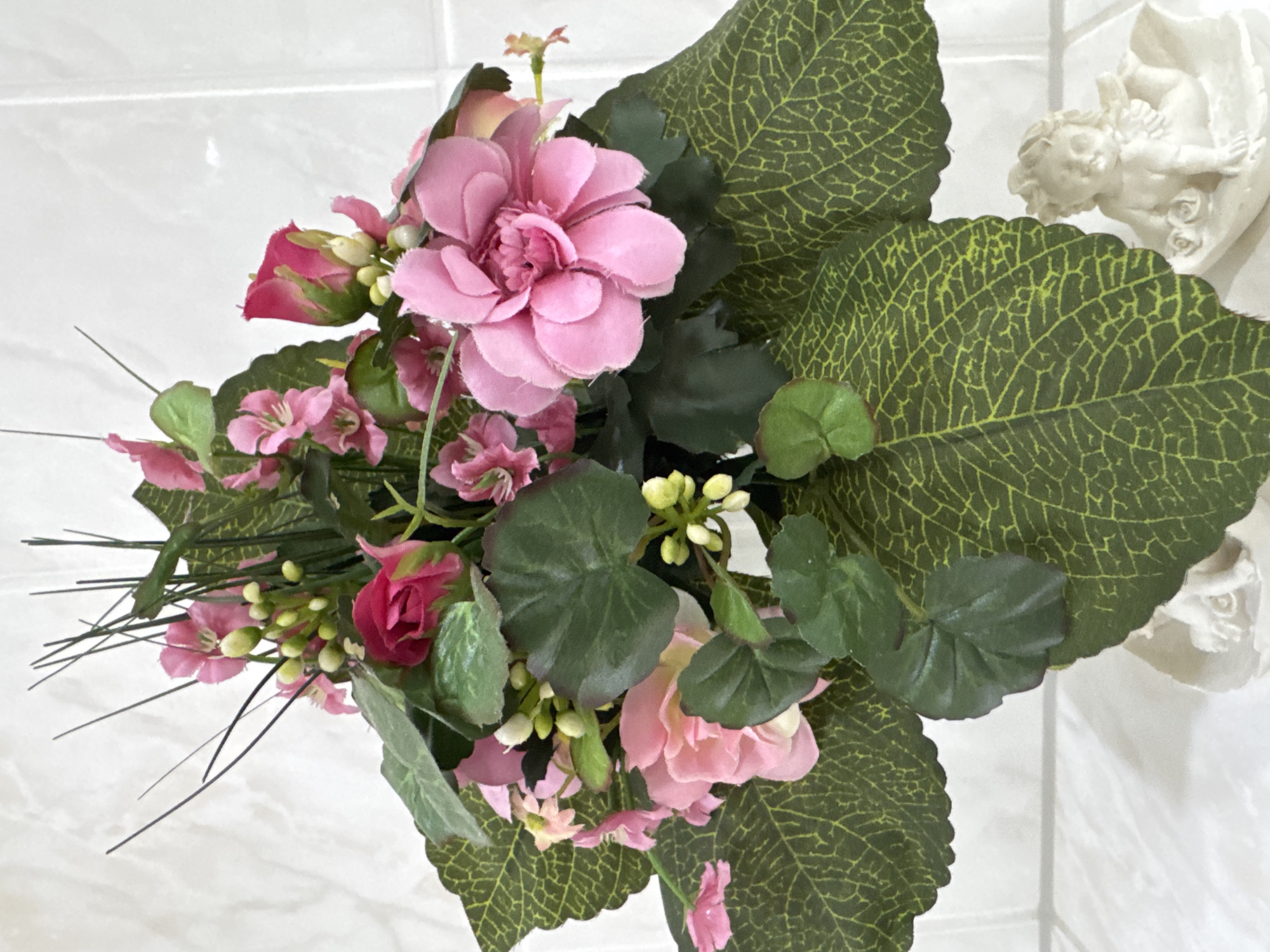 A still life photograph depicts a floral arrangement with pink and red flowers, green leaves, and thin, dark green sprigs. The flowers are a mix of roses and other blossoms with multiple petals. Some of the flowers are in full bloom, while others appear to be buds. The leaves are large and veined, with a textured appearance. In the lower right corner of the image, a small, white cherub figurine is visible, sitting on a decorative base with two roses. The background is a white tiled wall. The image is well-lit and the focus is on the floral arrangement and the cherub. The location is not specified in the image, but the scene is likely indoors, possibly in a home or a decorative setting within the city of Bietigheim-Bissingen, Germany.