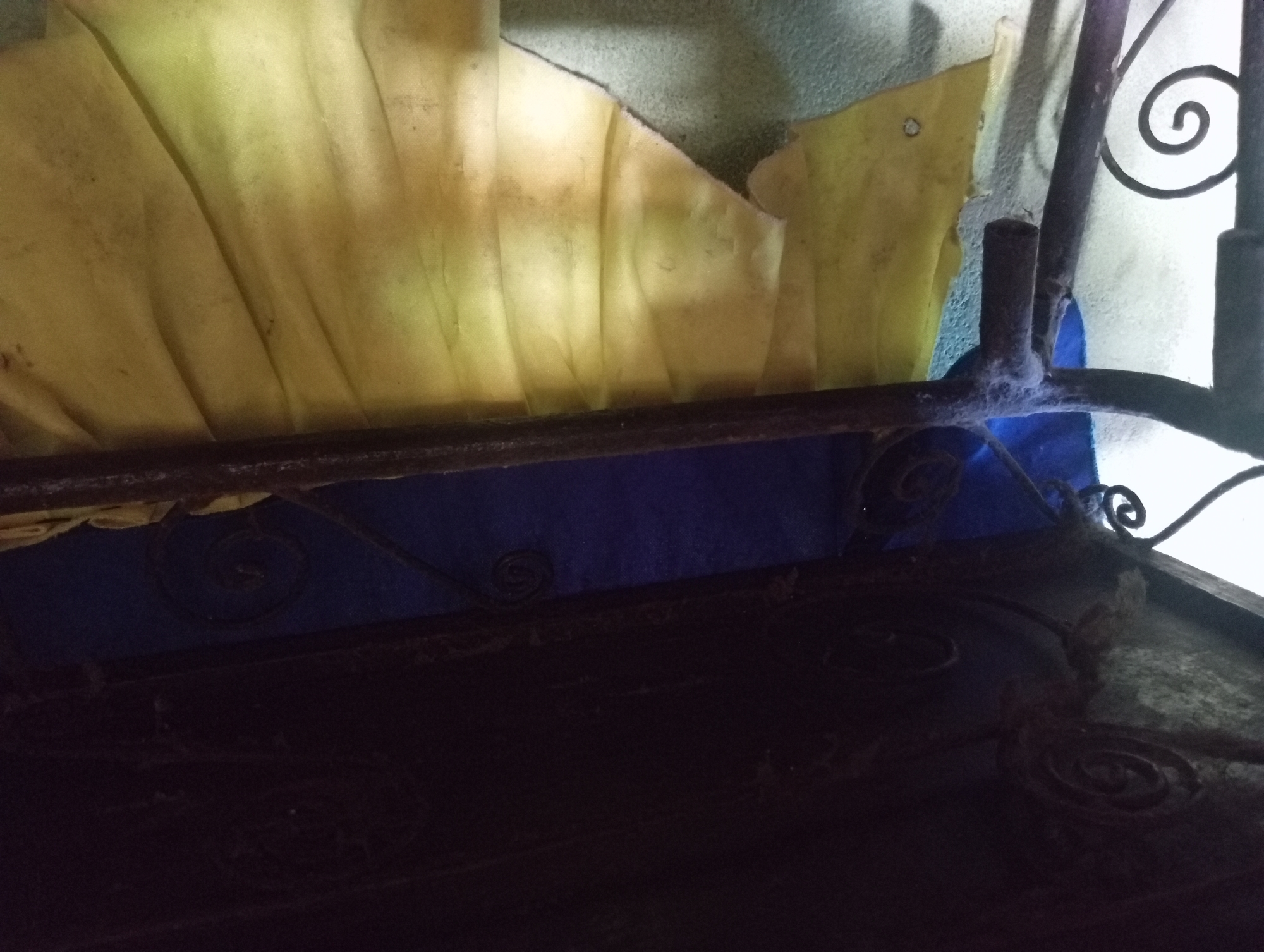 The image shows the underside of a dark, possibly metal, structure, perhaps a bed frame or a shelf. Decorative spiral metalwork is visible on the underside of this structure. Below this, there is a blue fabric or wall. Hanging down in front of the blue material is a crumpled yellow cloth or curtain, which appears dusty and possibly stained. Some part of a black metal frame with a spiral decoration is visible on the left side of the image. The background wall appears to be a textured, light-colored surface. The scene is dimly lit. The specific location in Rejosari, Indonesia, cannot be determined from the visual information present in the image.