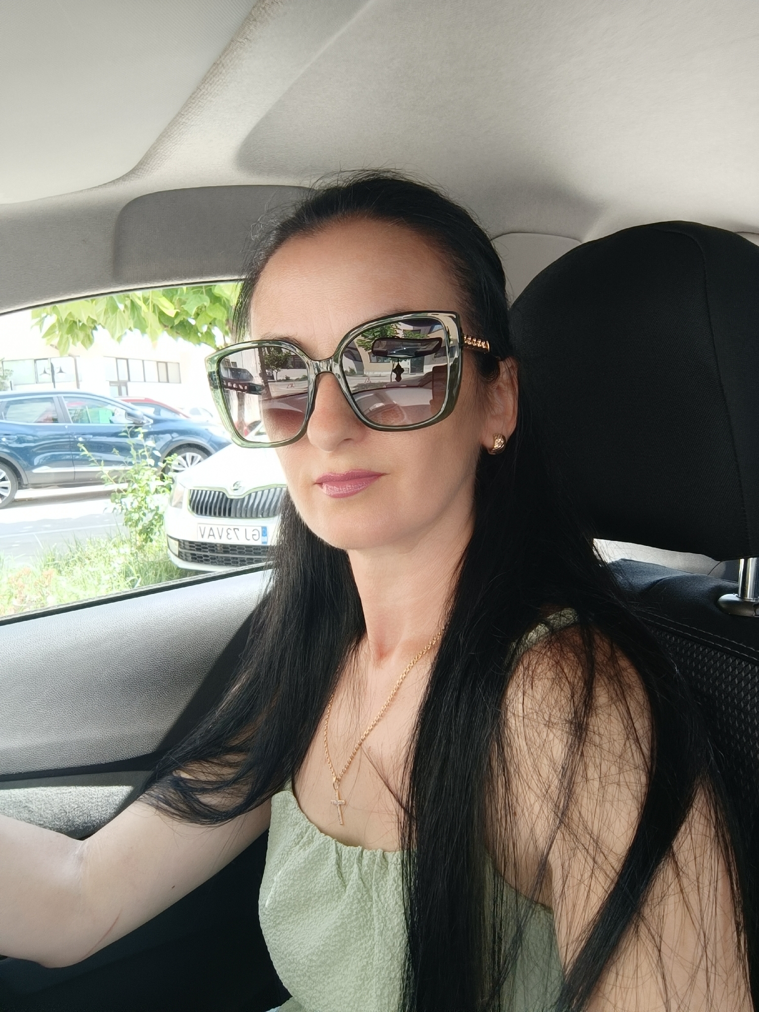 A woman with long, dark hair and large, dark-rimmed sunglasses is sitting in the driver's seat of a car. She is wearing a light green, textured top and a delicate gold necklace with a cross pendant. The interior of the car is visible, including the car's headrest and the passenger side door. Outside the car, through the window, other vehicles and buildings are visible, suggesting an urban environment. The license plate of the car in front of her appears to be in Romanian. The scene suggests a woman is about to depart or has recently arrived at a location in Târgu Jiu, Romania.