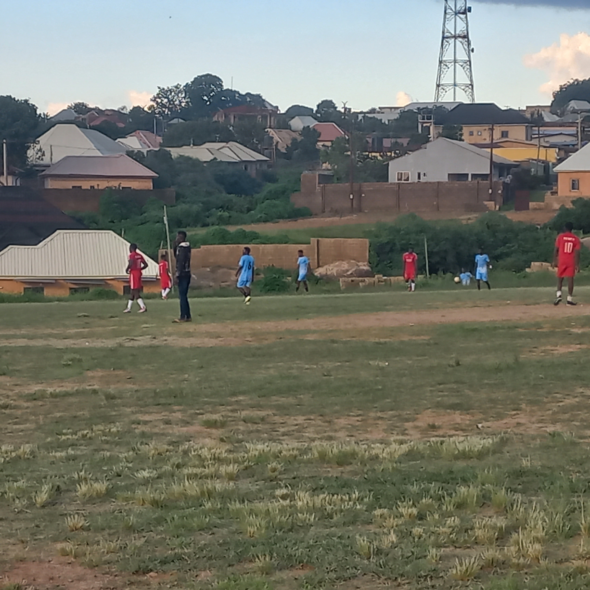 The image captures a group of young men playing soccer on a grassy field in Jos, Nigeria. In the background, a collection of residential buildings with diverse architectural styles, ranging from modern to traditional, are visible. Some of these structures are painted in earthy tones, while others feature white or grey facades, indicative of the varied development in the area. A prominent telecommunications tower stands tall against the sky, a common landmark in urban and semi-urban Nigerian settings. The sky displays a mix of blue and scattered clouds, suggesting it might be late afternoon or early evening. The overall scene depicts a typical day in a residential area of Jos, where recreational activities like soccer are common amongst the youth.