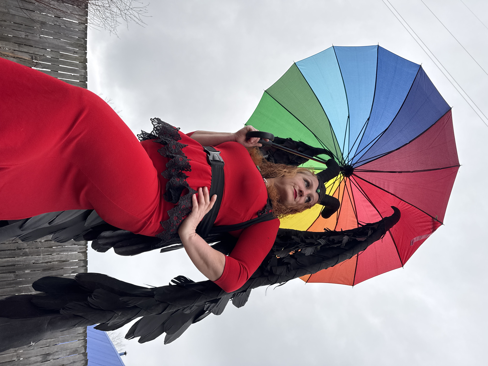 The image shows a woman dressed in a red dress with black lace and feathered black wings. She is holding a rainbow-colored umbrella and wearing horns on her head. She appears to be at an outdoor location in the city of Chaikovsky, Russia, evidenced by the backdrop of a cloudy sky and surrounding buildings.