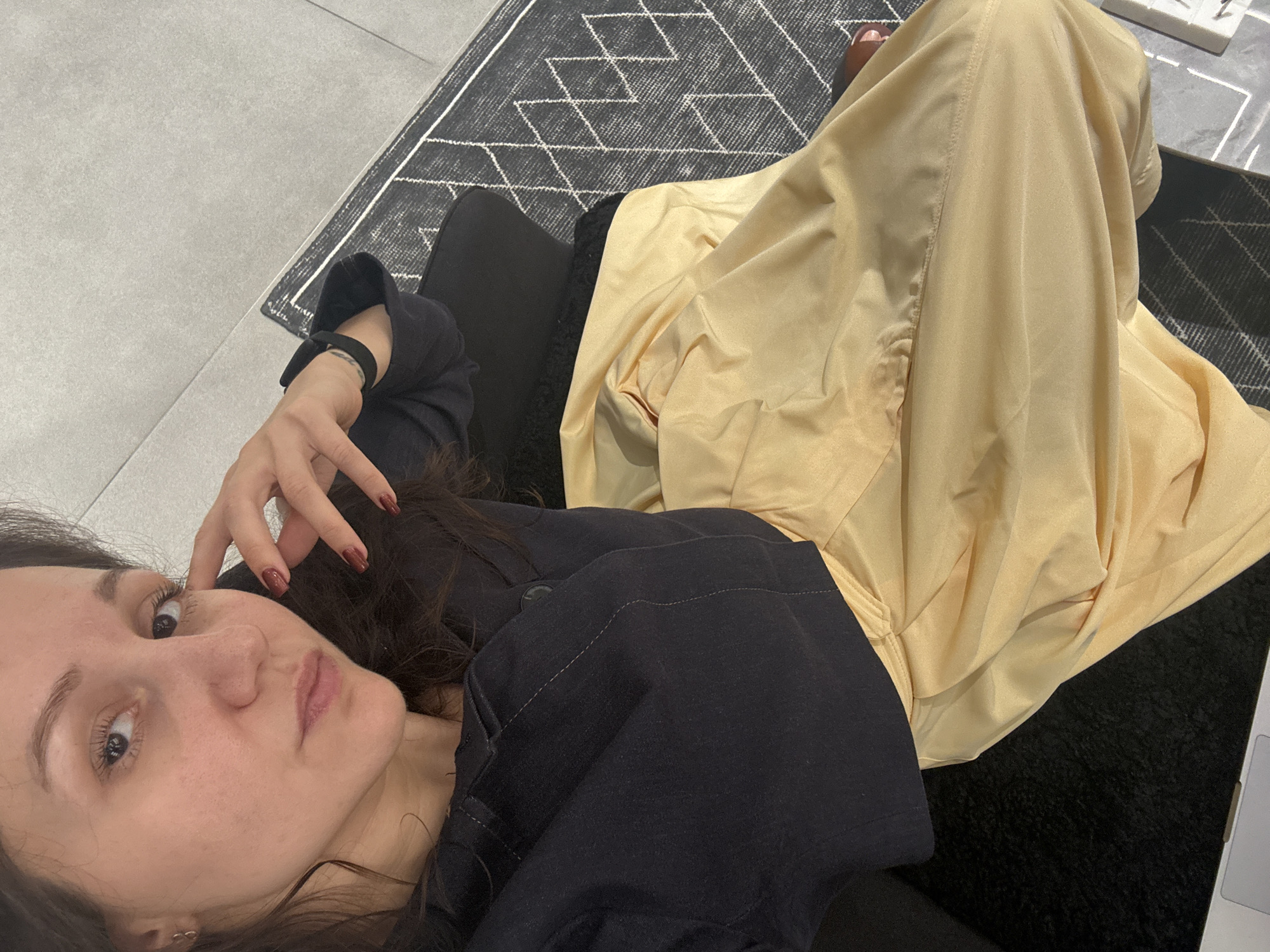 A woman with fair skin, brown hair, and dark red nail polish is lying on a dark-colored couch. She is wearing a dark-colored top. The woman is looking at the camera. She is wearing a black fitness tracker on her left wrist. The woman is partially covered by a gold-colored, draped fabric. The couch is on top of a gray rug with a geometric pattern. The floor is gray, and there are white objects in the top right corner. The location is unknown and there is no evidence suggesting it is in Dubai, United Arab Emirates.