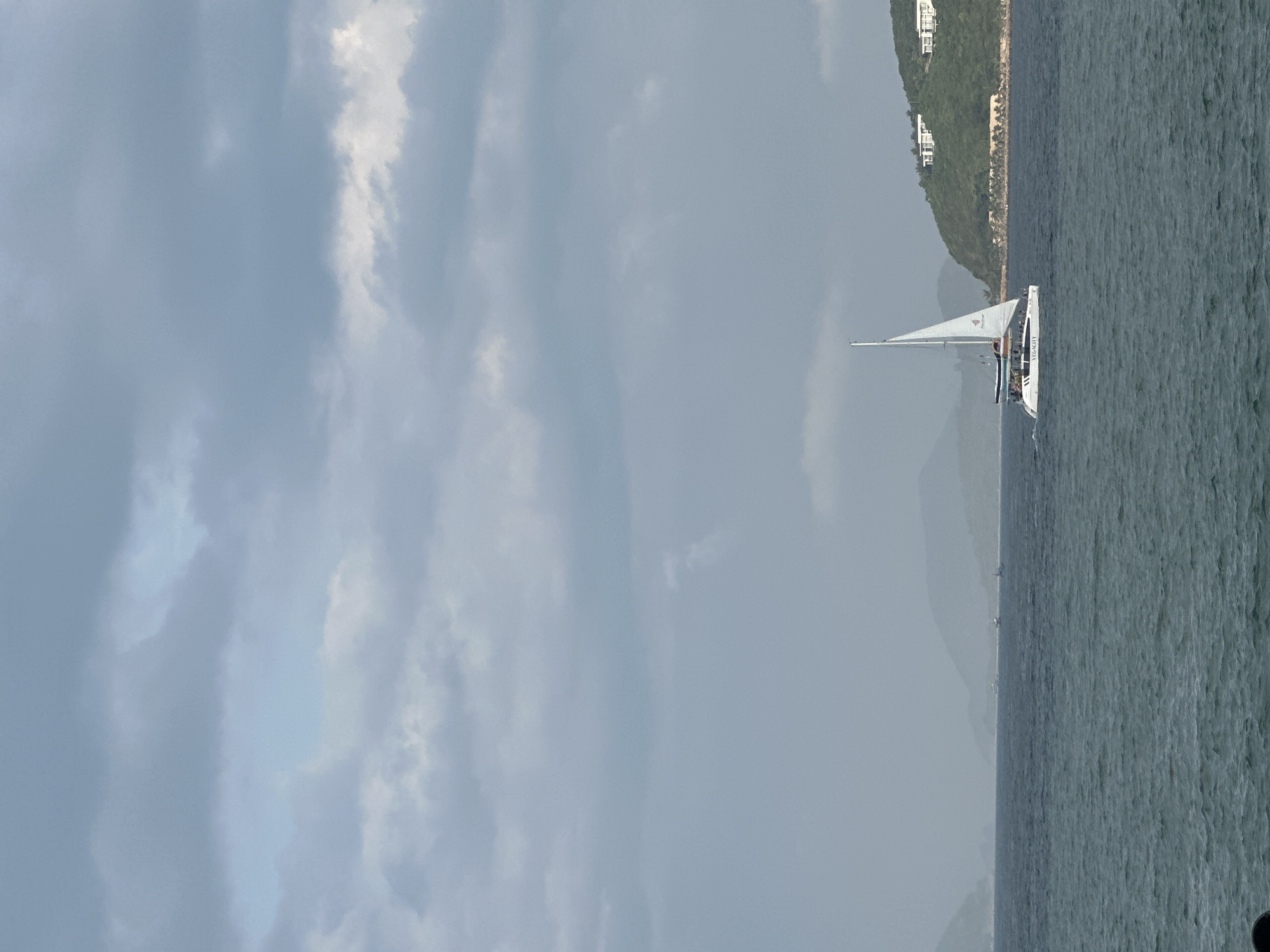 This image depicts a seascape within Nha Trang Ward, Vietnam, under an overcast sky. The upper two-thirds of the frame are dominated by a dense, grey-blue cloud cover with patches of lighter sky visible. The lower third of the image shows a choppy, dark blue-green body of water.
In the mid-ground, a white catamaran named "VEGACITY" is sailing from left to right. It has a single mast with a white mainsail bearing a red logo. Several indistinct figures, consistent with passengers, are visible on the catamaran's deck. Behind the catamaran, a small wake is observable.
To the right, a verdant, tree-covered hillside rises from the water, topped by two distinct white, multi-level modern buildings. In the far distance, multiple layers of mountains or hills appear as faint, hazy blue-grey silhouettes. Several small, dark vessels are visible on the water's surface in the distance, against these remote landmasses. The overall lighting is diffused, indicative of a cloudy day.