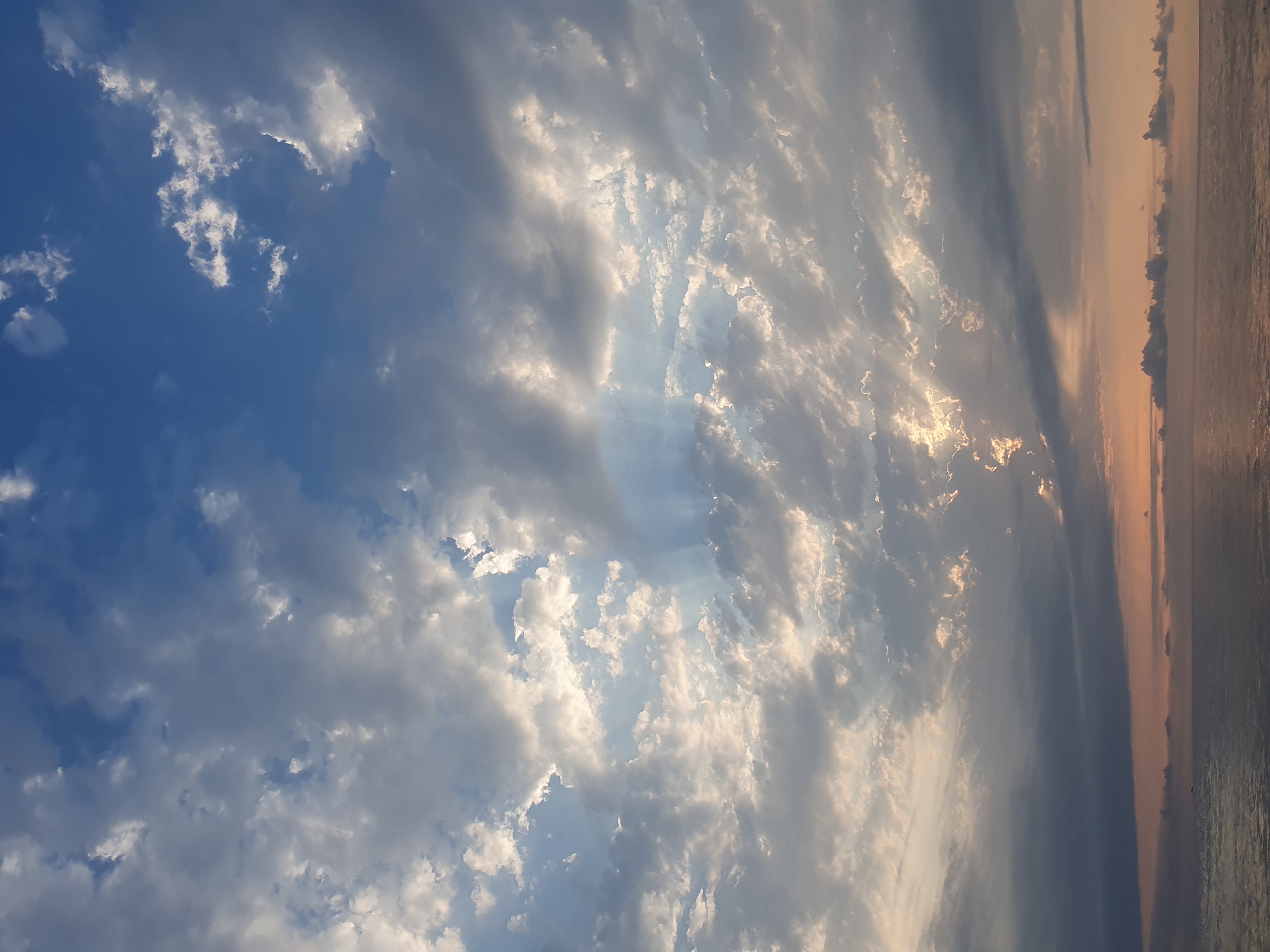 The image depicts a cloudy sky over the sea, with rays of sunlight piercing through the clouds. The lower portion of the image shows the calm, reflective surface of the sea, with a soft, diffused light emanating from the horizon. The sky is a mixture of blue and gray hues, with various cloud formations, some dense and dark, others thin and wispy. The sunlight creates dramatic streaks and illuminates patches of the clouds, giving the scene a serene and ethereal quality. The location is the coast of Batumi, Georgia. There are no people or man-made objects visible in the frame.