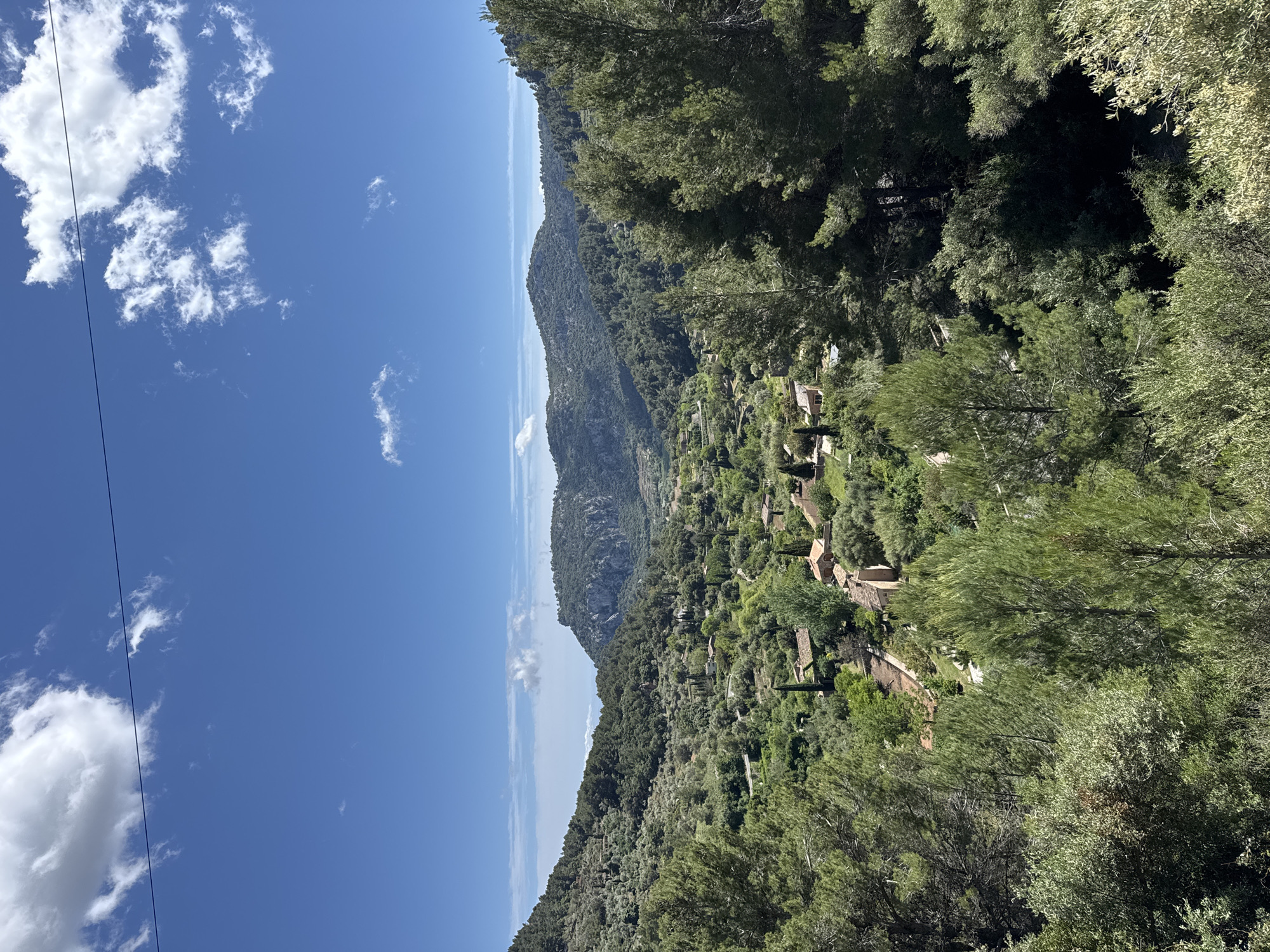The image shows a mountainous landscape in S'Arxiduc, Spain. The sky is blue with scattered white clouds. A mountain range runs along the horizon. The foreground is dominated by lush green trees and vegetation. Small buildings are nestled amongst the trees. A utility wire stretches across the top left of the image.