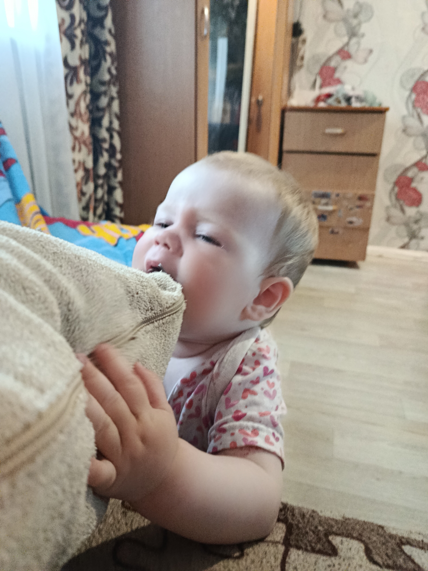 A baby with fair skin and blonde hair is biting the corner of a tan-colored blanket. The baby is wearing a long-sleeved shirt with a pink heart pattern. One hand is holding the blanket. The background shows a room interior with wallpaper featuring a floral pattern in white and red, a wooden cabinet, curtains with a brown and beige print, and a floor with a dark brown rug. The location is Фоки, Russia.