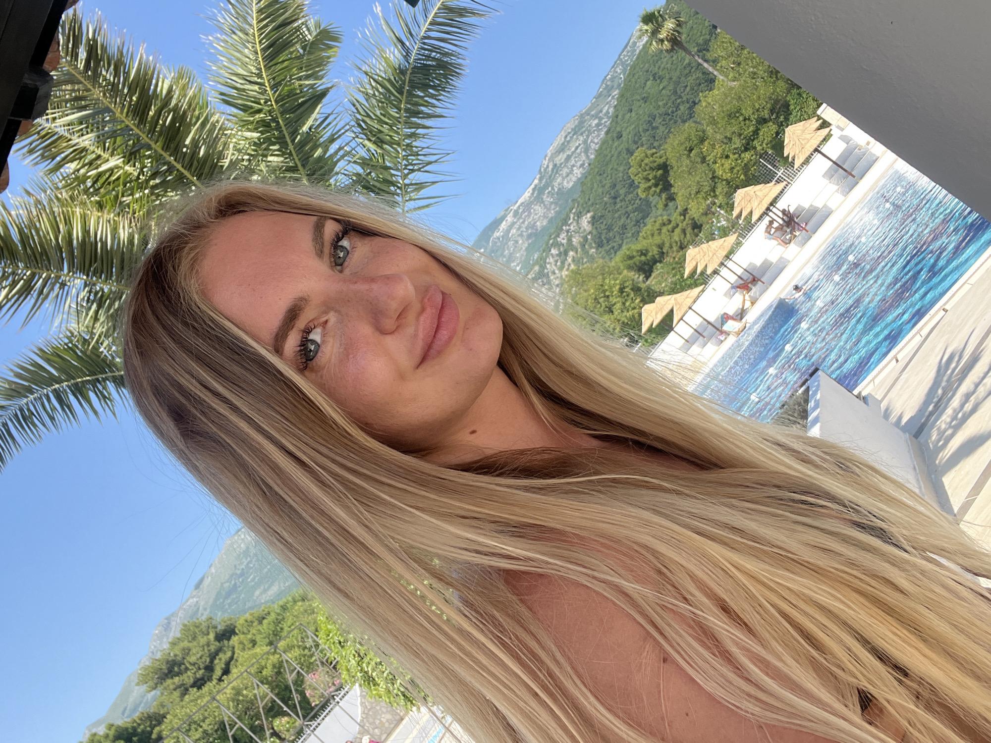 A medium-angle, outdoor selfie features a blonde woman in front of a swimming pool, a palm tree, and a mountain. The woman has fair skin, blue eyes, and wears subtle makeup. Her long, blonde hair is styled straight and falls over her shoulder. She is looking at the camera with a slight smile. Behind her, there's a blue swimming pool with people sunbathing on lounge chairs under straw umbrellas. In the background, a green, mountainous landscape is visible under a clear blue sky, suggesting the location is Budva, Montenegro. A corner of the building is visible at the top left of the image.