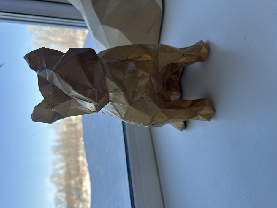 A low-poly golden animal, possibly a bear, is mounted on a white wall near a window. The window reflects a clear blue sky. The geometric facets of the sculpture create an angular, modern aesthetic. Part of another similar, but lighter-colored, geometric sculpture is also visible above the golden one. Both pieces appear to be constructed from paper or a similar lightweight material.
