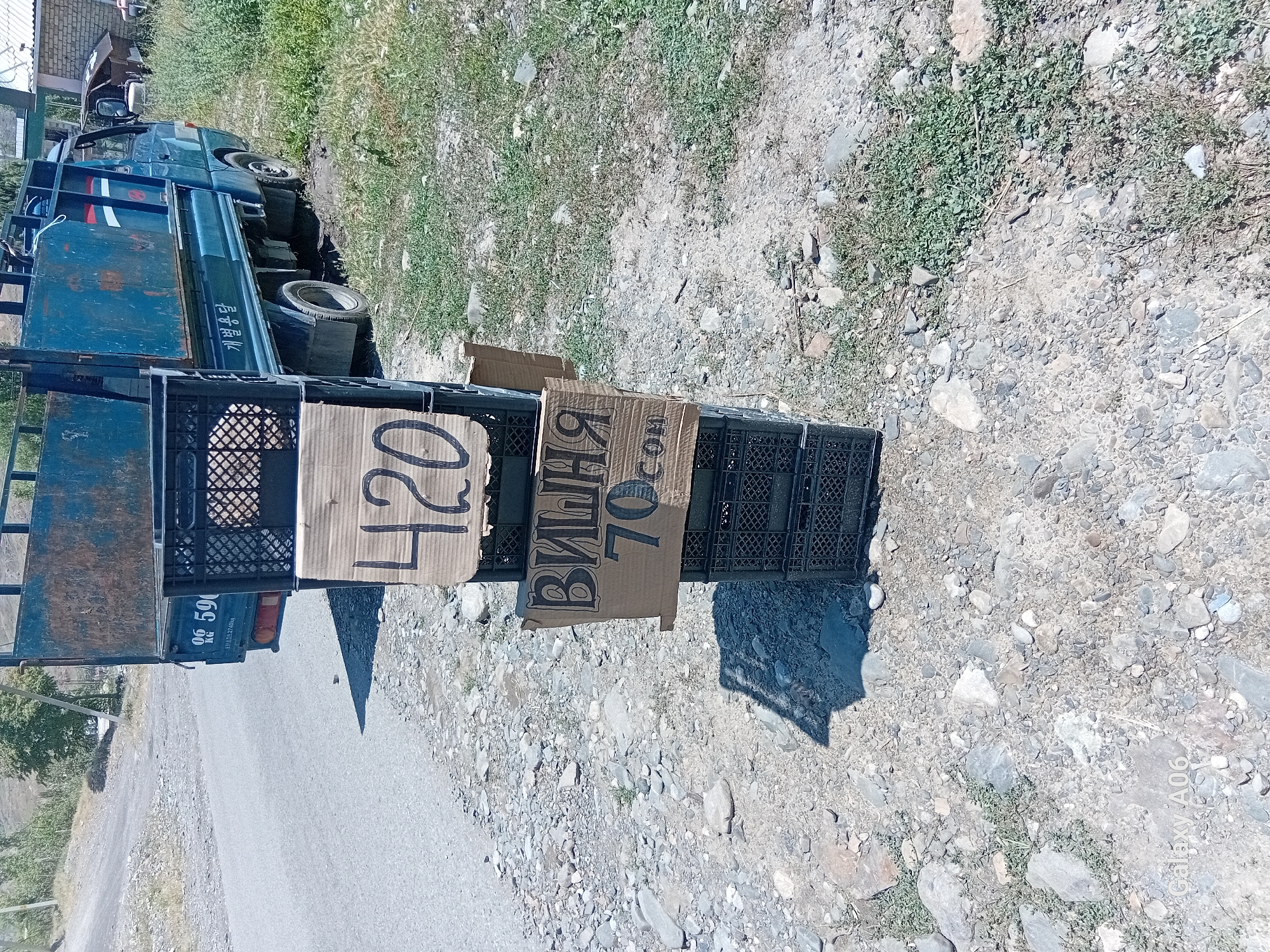 The image shows a blue truck parked on a dirt road, likely in Kojo-Aryk, Kyrgyzstan. The truck is an older model with a flatbed. Next to the truck, several black plastic crates are stacked, and two pieces of cardboard with writing on them are placed on top of the crates. The cardboard on the left has "420" written in black marker. The cardboard on the right has Cyrillic text and numbers, reading "ВИШНЯ 70 .com". The ground around the truck is covered with gravel and dry grass, suggesting a sunny and dry day. The truck has some signage on its side, with Korean characters and numbers. The overall scene appears to be an informal roadside sale or perhaps a vendor preparing to sell goods. The lighting suggests it is daytime, likely late morning or afternoon.