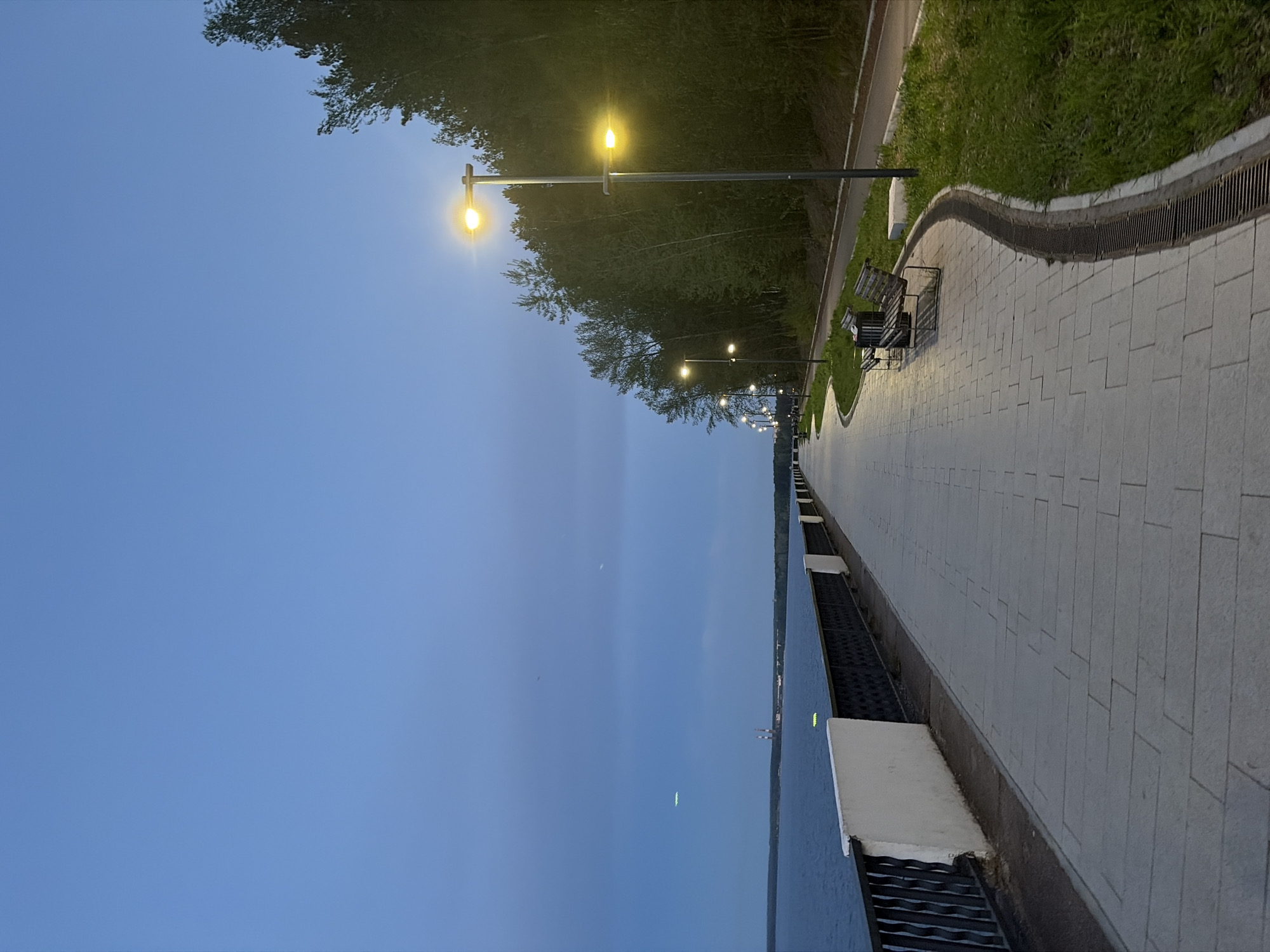 The image depicts an evening scene in the city of Chaikovsky, Russia. A paved embankment stretches along a body of water, likely the Kama River. Streetlights illuminate the walkway, and benches are placed along the edge of the path. Lush greenery lines the other side of the embankment. The sky is a dusky blue. The scene appears serene and still.
